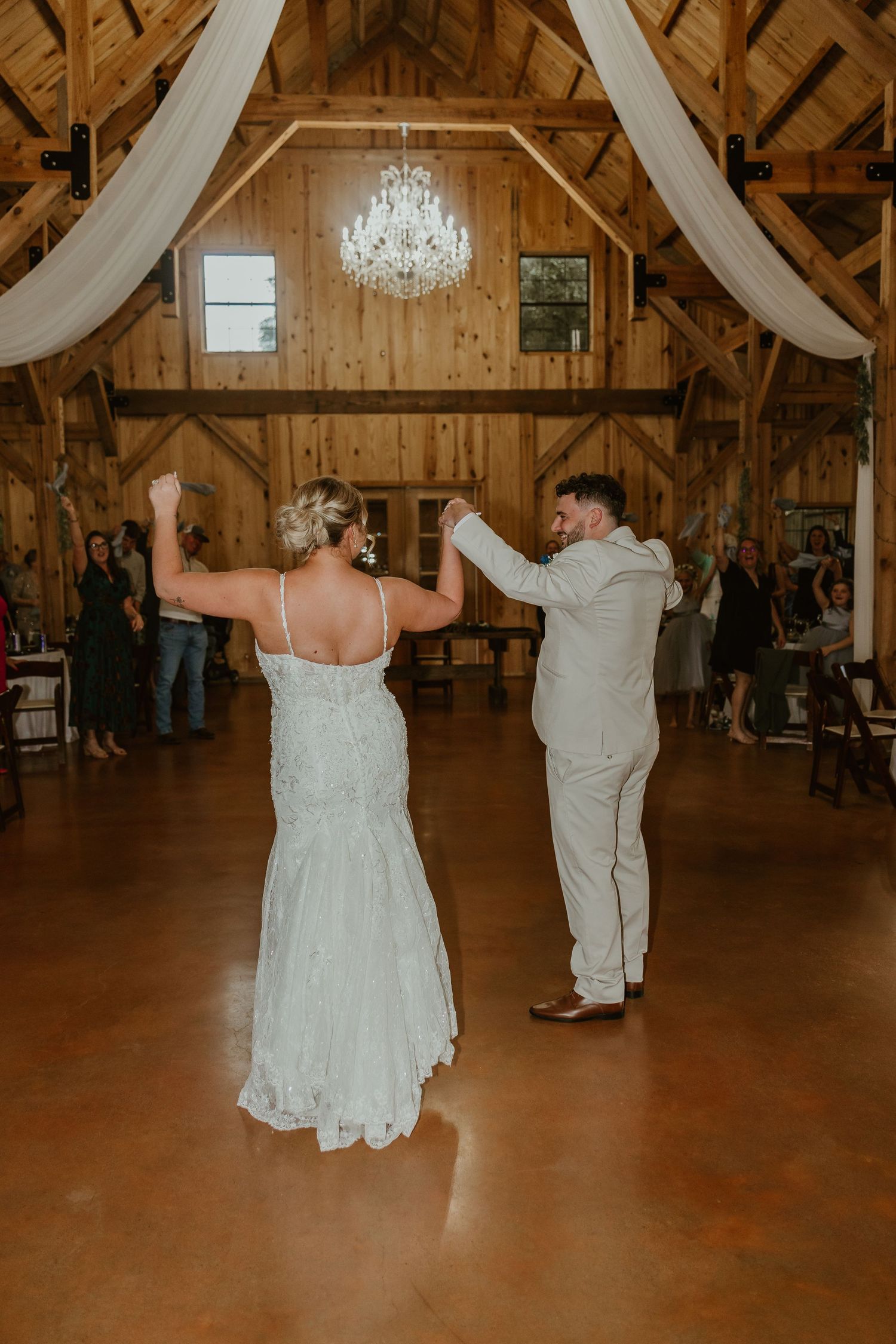 A couple dances together in a rustic barn venue with elegant chandeliers and white draping.