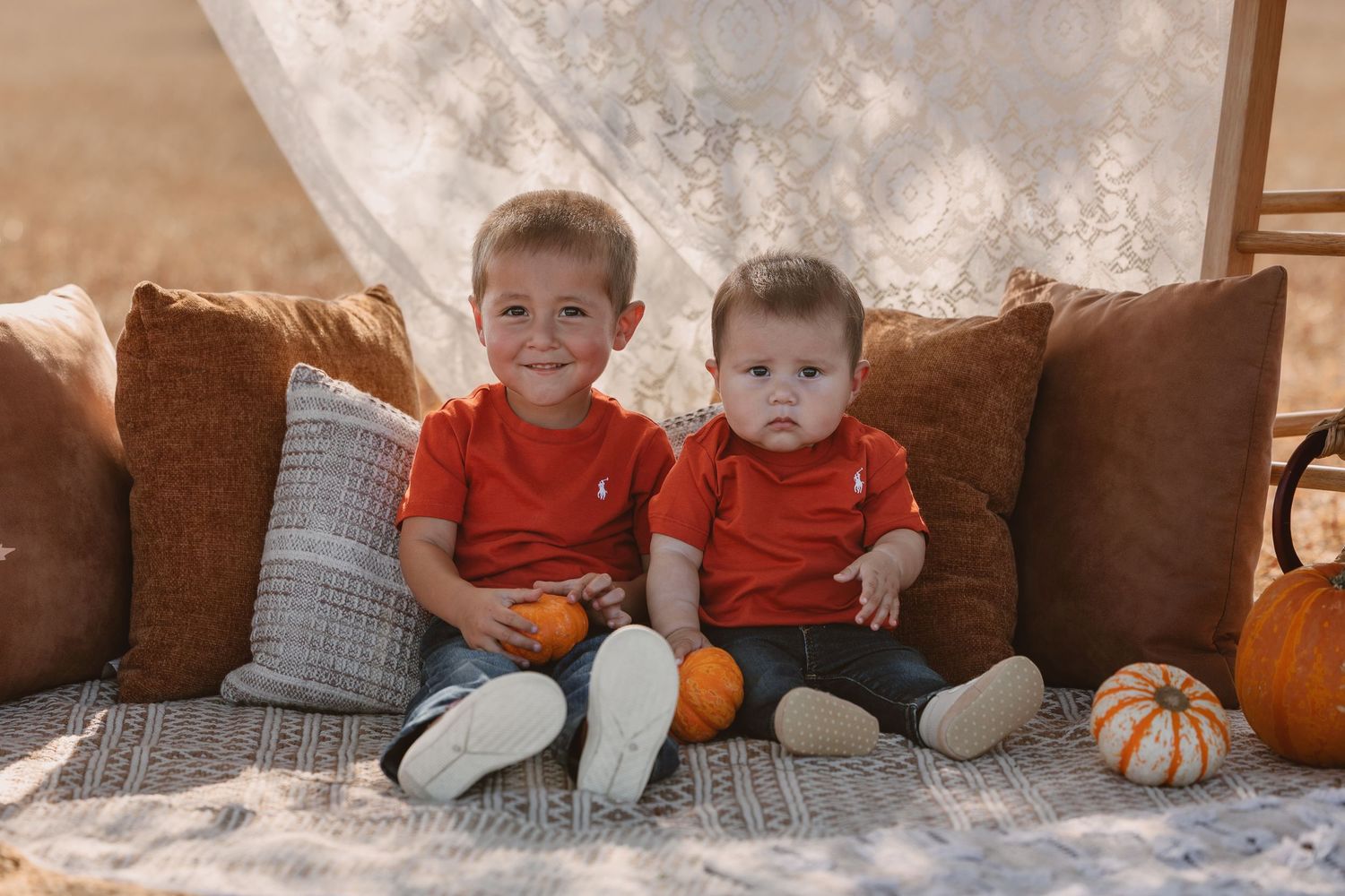 Two young brothers in matching red shirts sit together surrounded by pumpkins in a fall photo session.