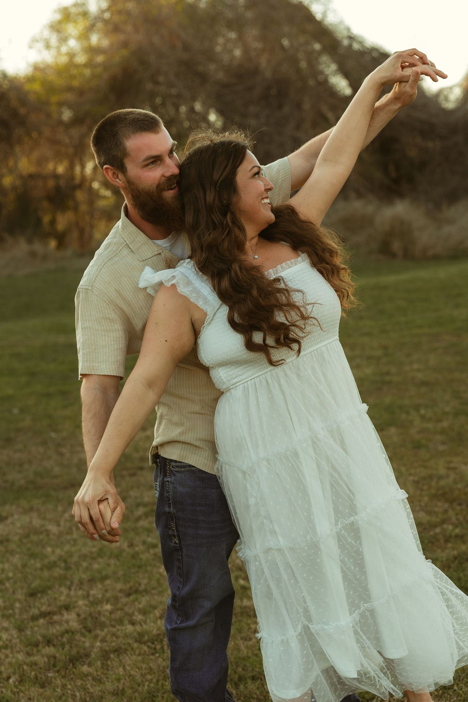 A couple dances outdoors at golden hour, woman in white dress with arm raised, man in beige shirt and jeans smiling.