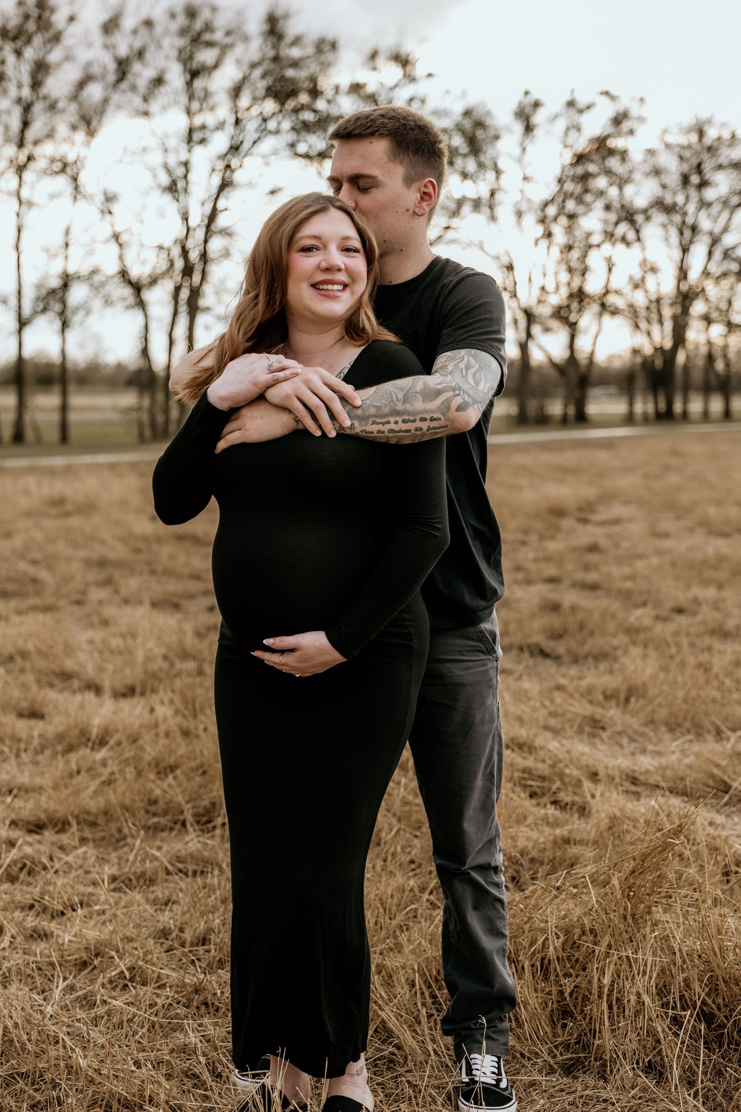 An expecting couple shares a tender moment in a field at sunset.