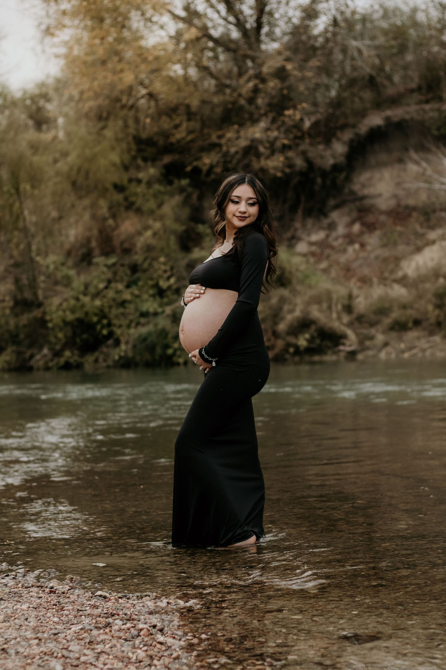 Expectant mother poses gracefully in flowing black dress by river's edge.