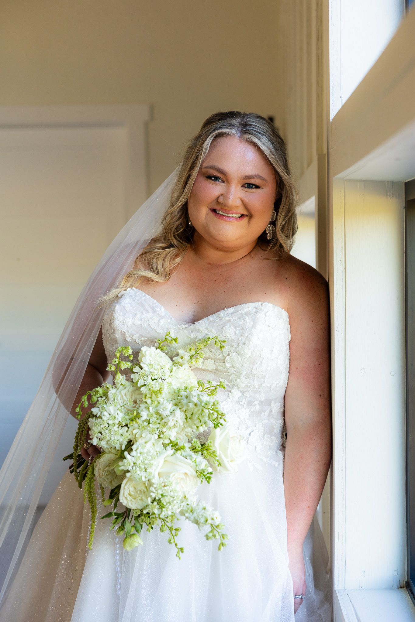 Smiling bride in strapless lace gown with veil holding white and green cascading bouquet by a sunlit window.