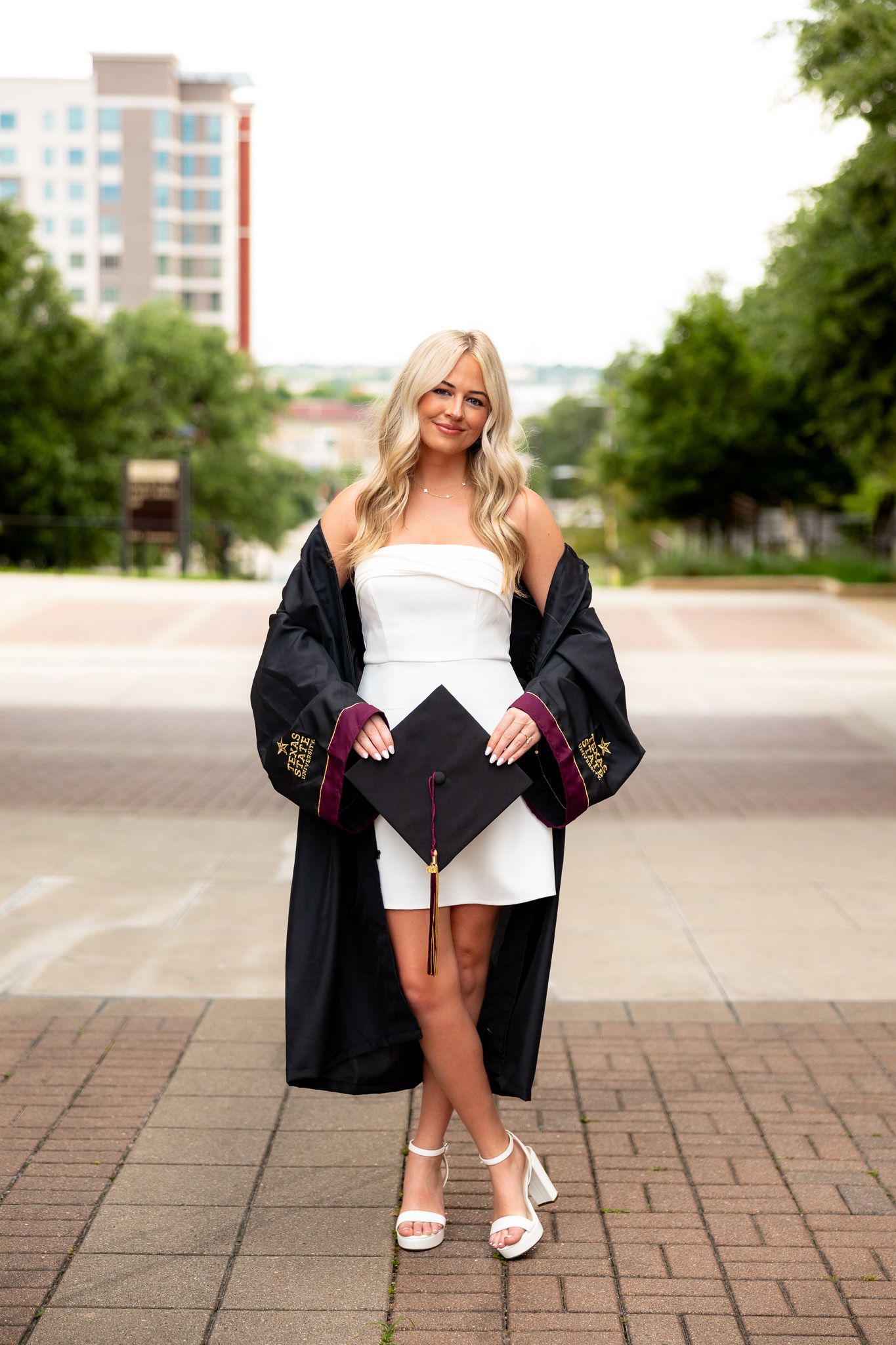 Smiling blonde graduate in white dress holding cap, wearing black graduation gown, posing outdoors on a brick walkway.