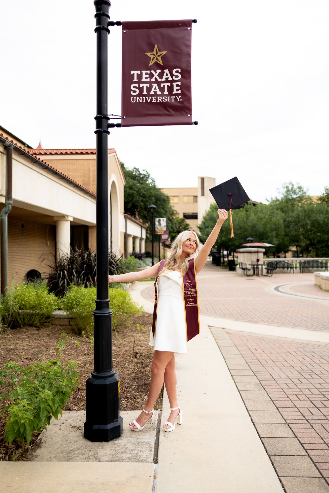 A smiling female graduate in white dress holds cap aloft beside Texas State University banner on campus sidewalk.