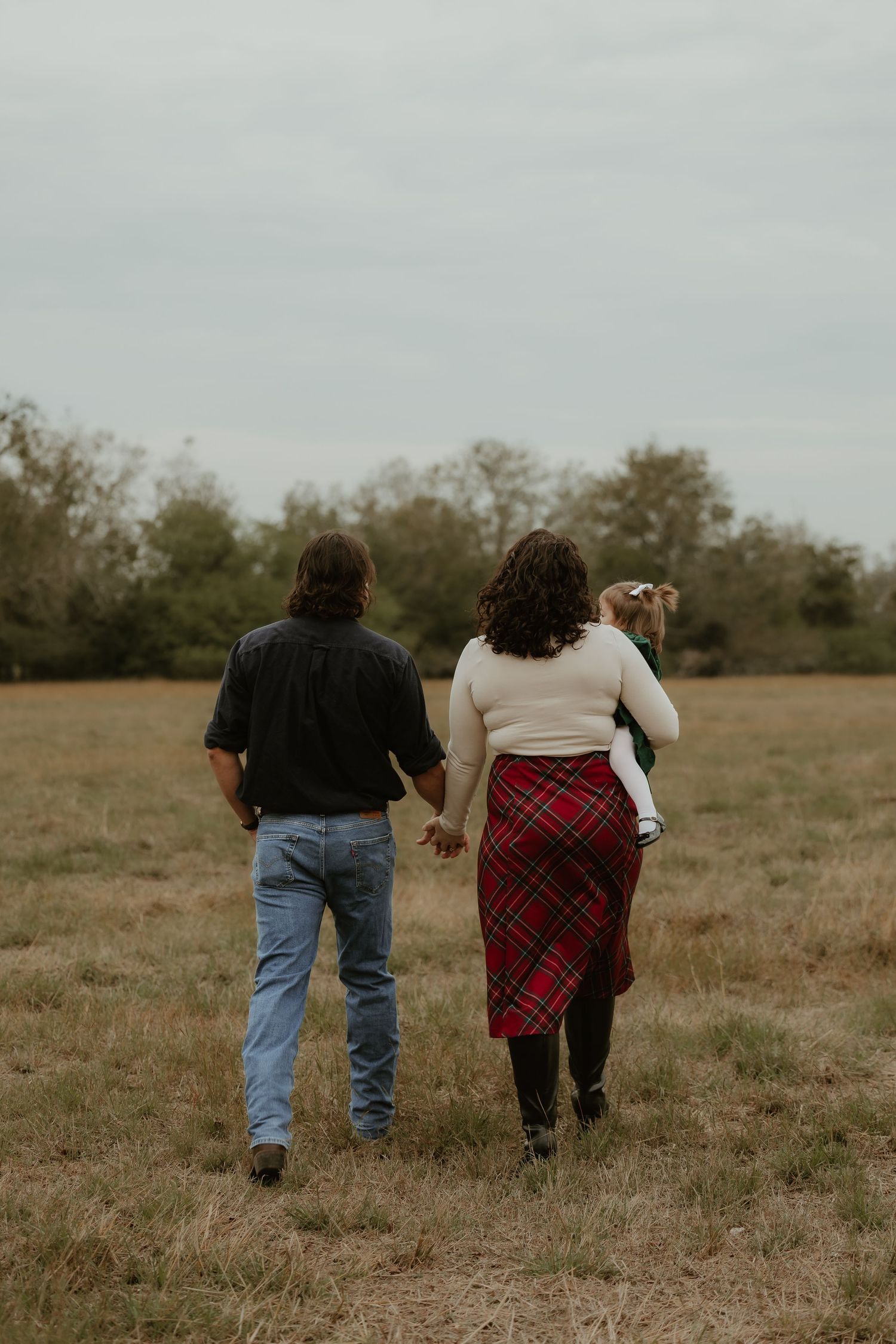 A family walks hand-in-hand through an open field, woman in red plaid skirt carrying a child, man in jeans and black shirt.