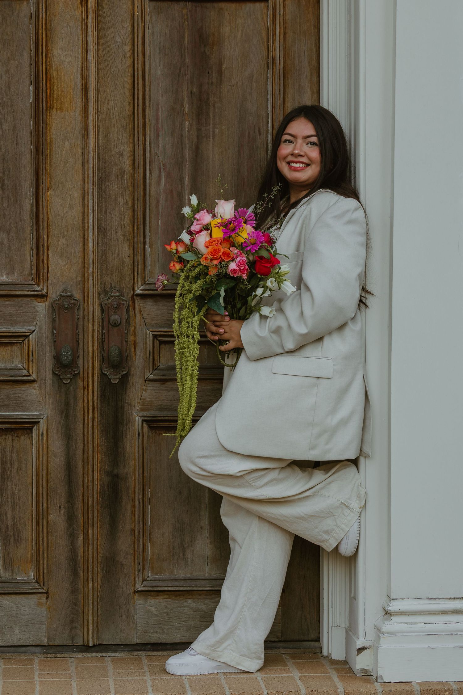 Stylish poses by wooden door featuring white suit and colorful flower arrangement.