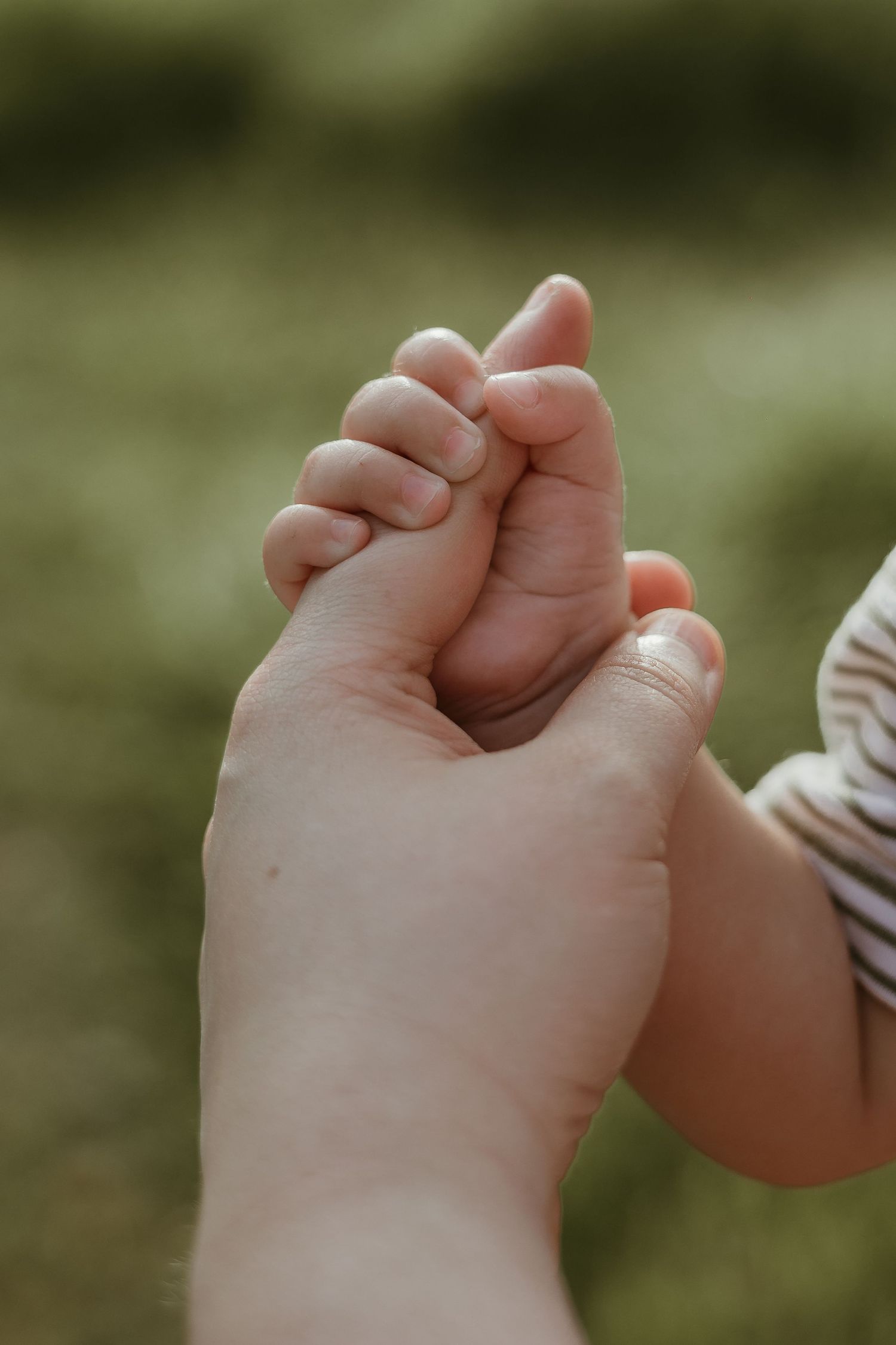 Close-up of tiny baby hand holding mom's finger against blurred green background.