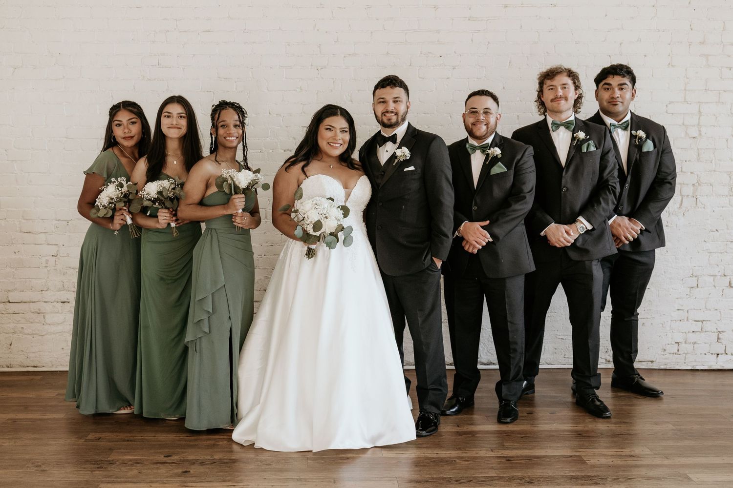 Full wedding party poses in formal black suits and sage green dresses.