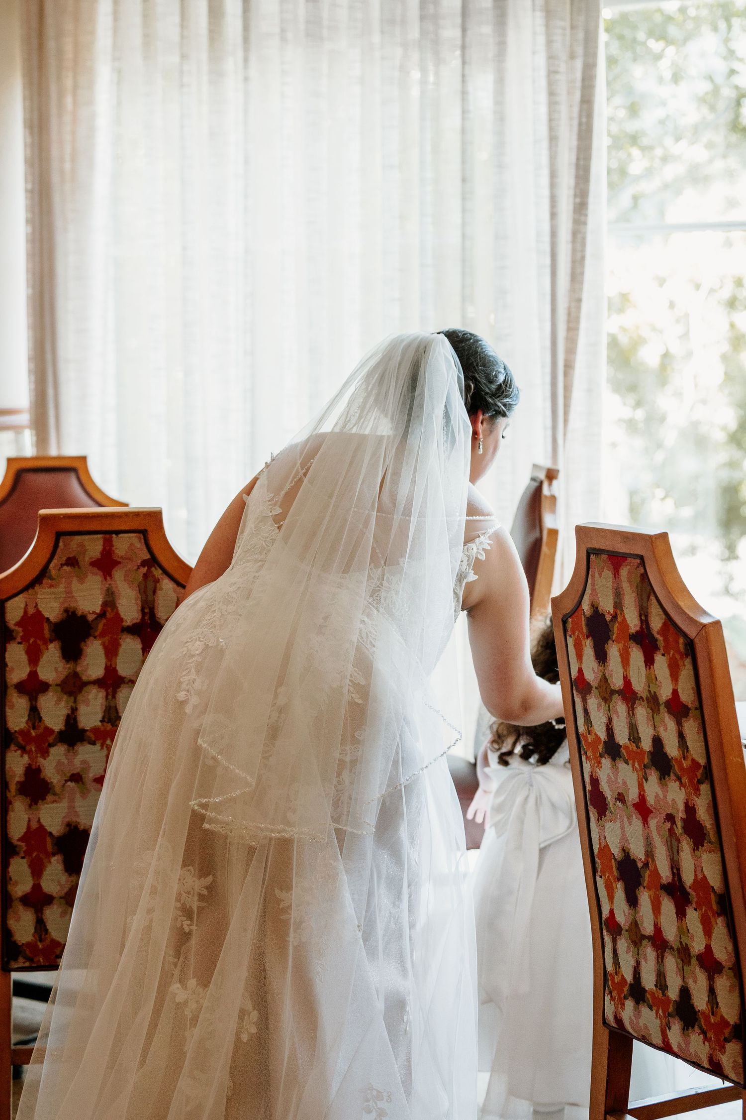 A bride in a flowing veil stands by ornate patterned chairs near a window.