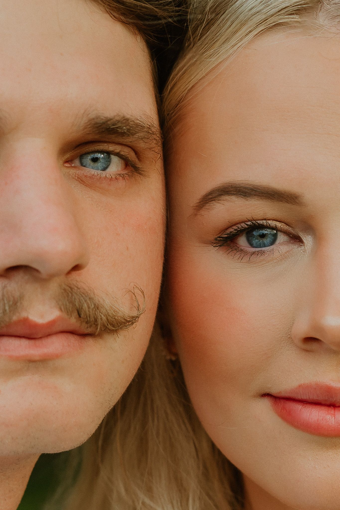 Close-up portrait of a man with a mustache and a blonde woman, both with blue eyes, cheeks touching.