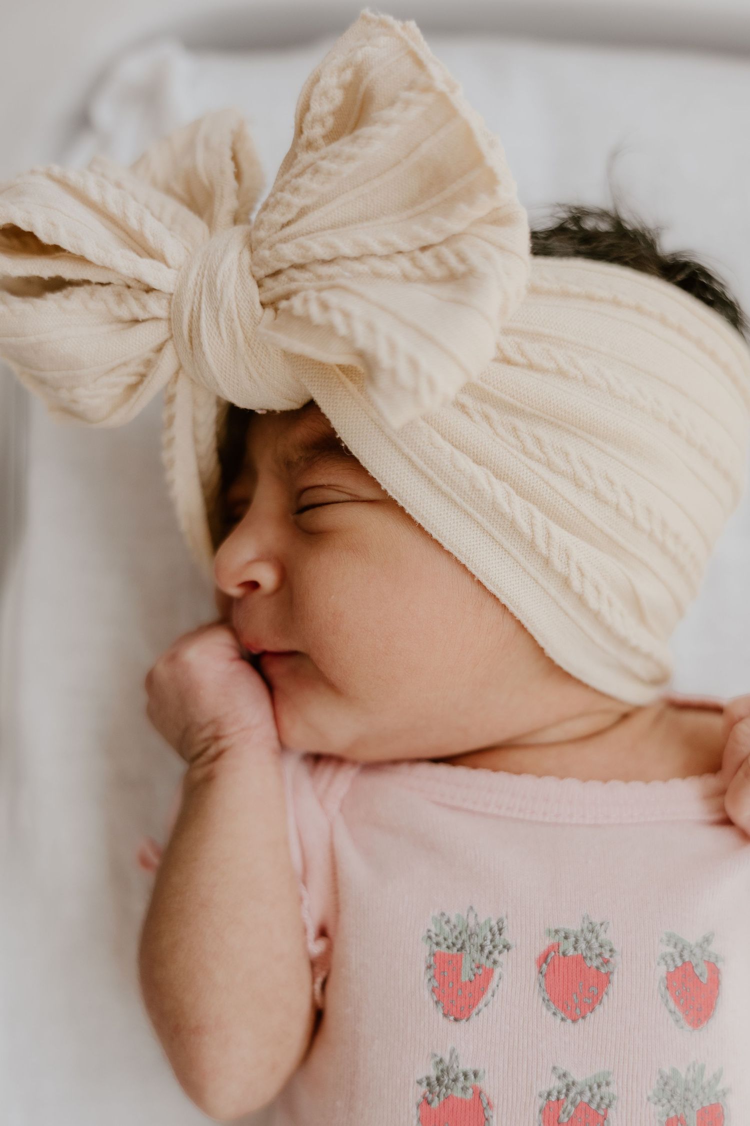 Sleeping newborn baby wearing a cream bow headband and pink strawberry onesie.
