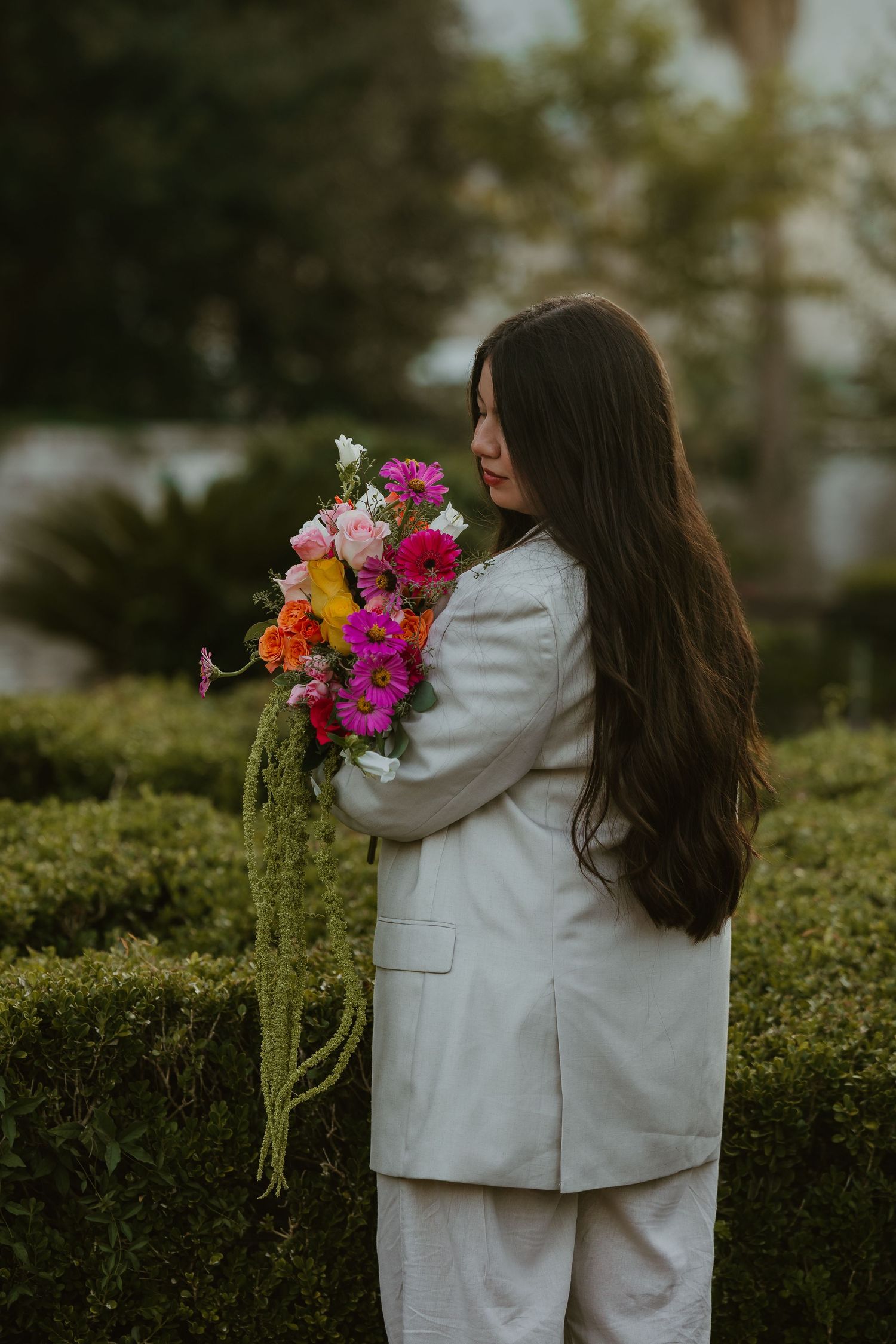 Woman in white suit holding vibrant pink and orange flower bouquet, standing in front of manicured green hedges.