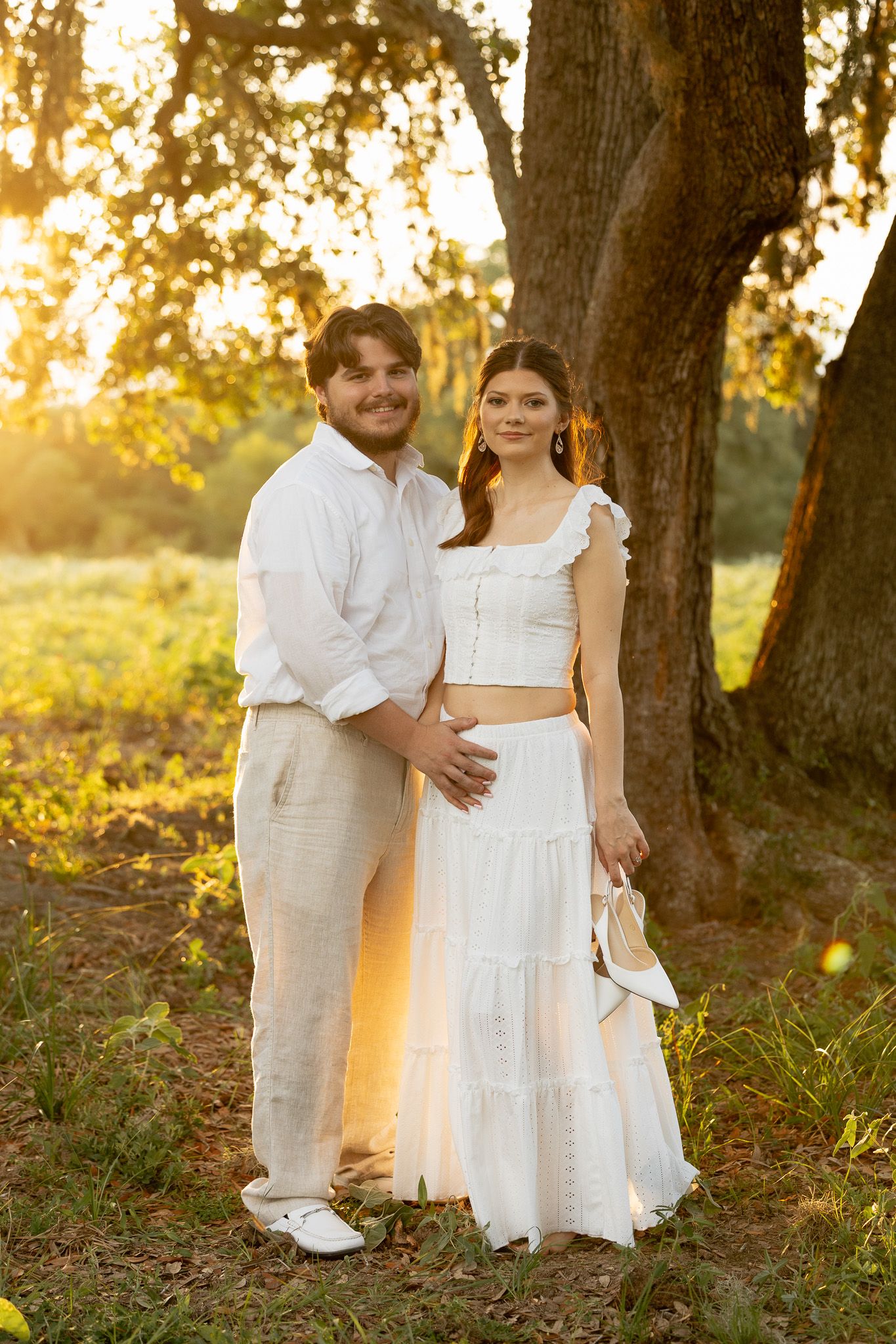 Young couple in matching white outfits posing together under a large oak tree during golden hour sunset outdoors.
