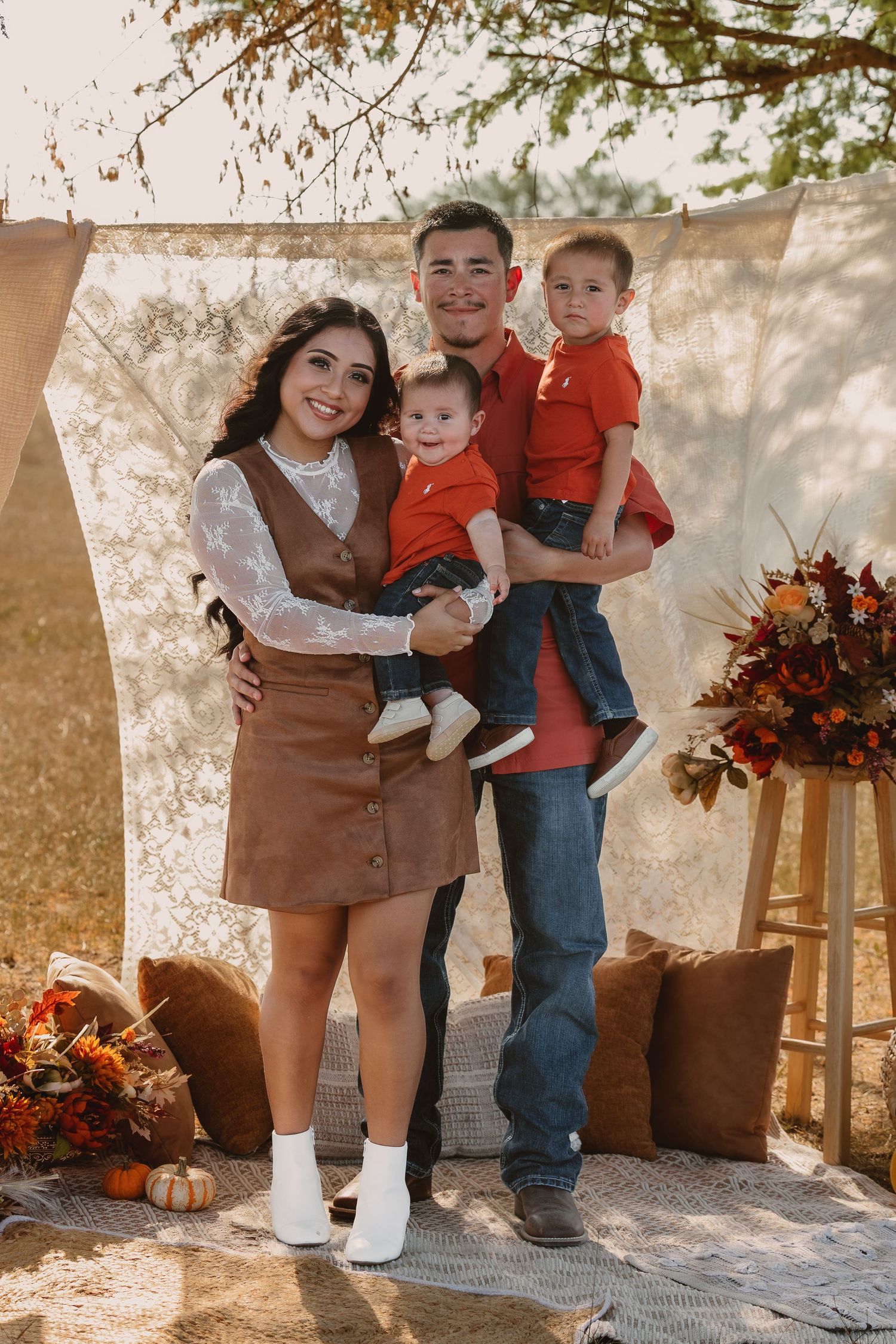 Family of four posing for a fall portrait outdoors, with two young boys in red shirts, autumn decor, and warm golden light.