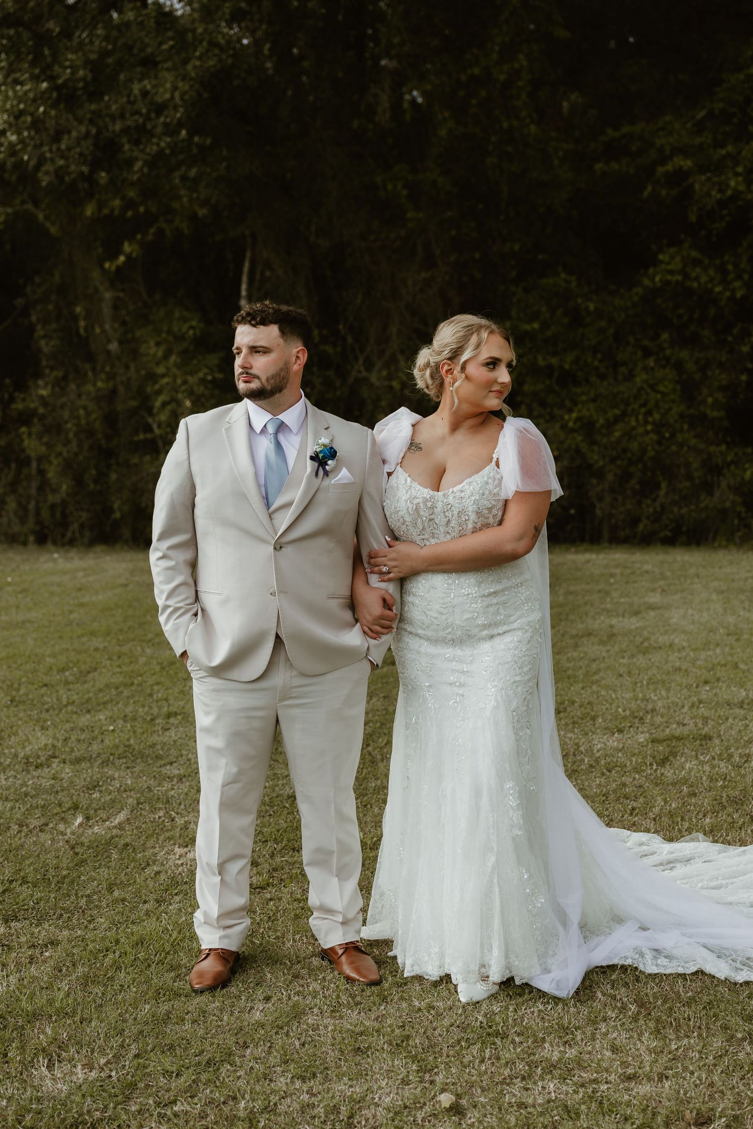 A couple in wedding attire poses together on a grassy field with trees in the background.