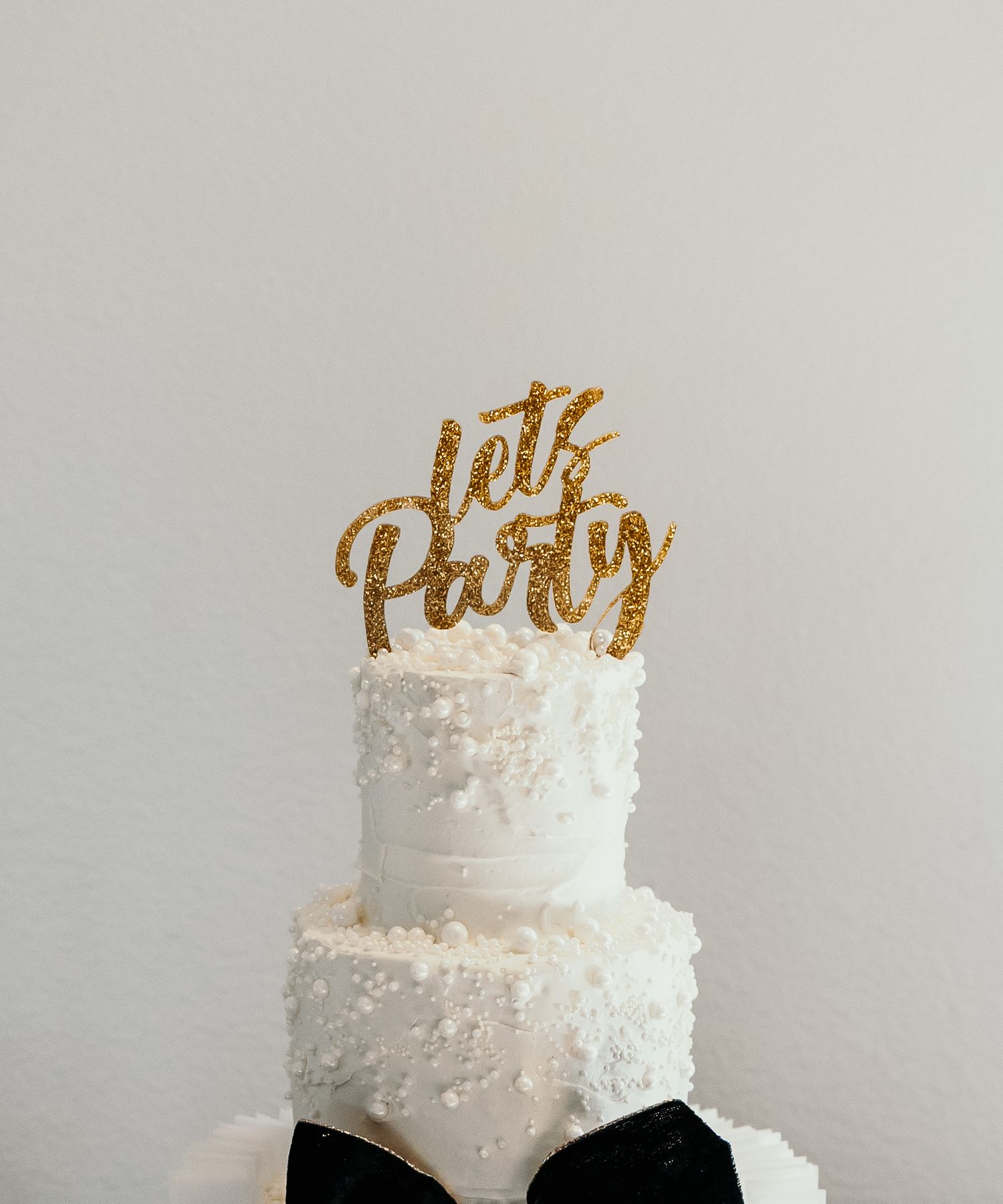 White wedding cake with gold glitter topper and black bow decorations displayed in pearls.