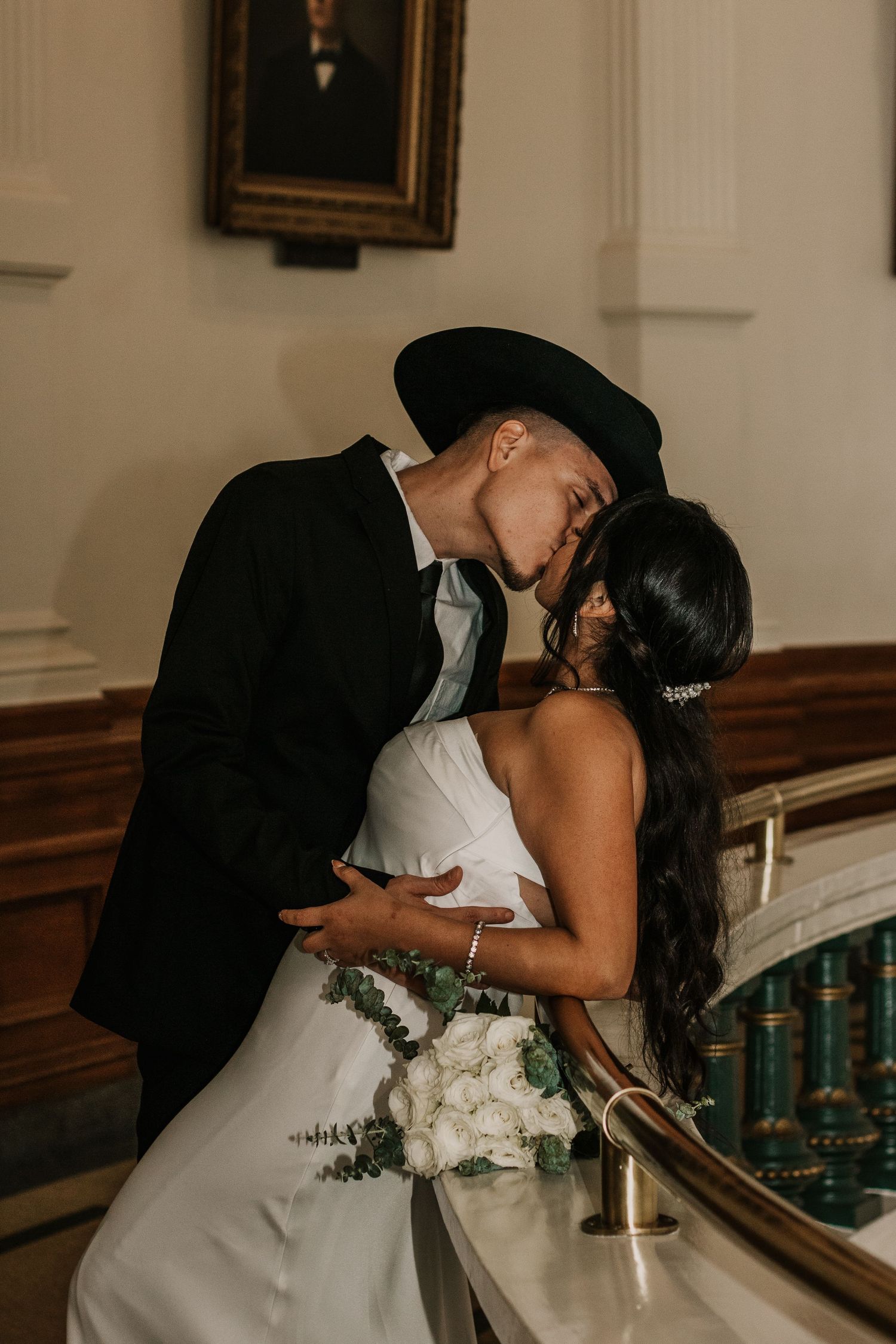 A couple shares a romantic kiss in formal attire beside an ornate indoor staircase.