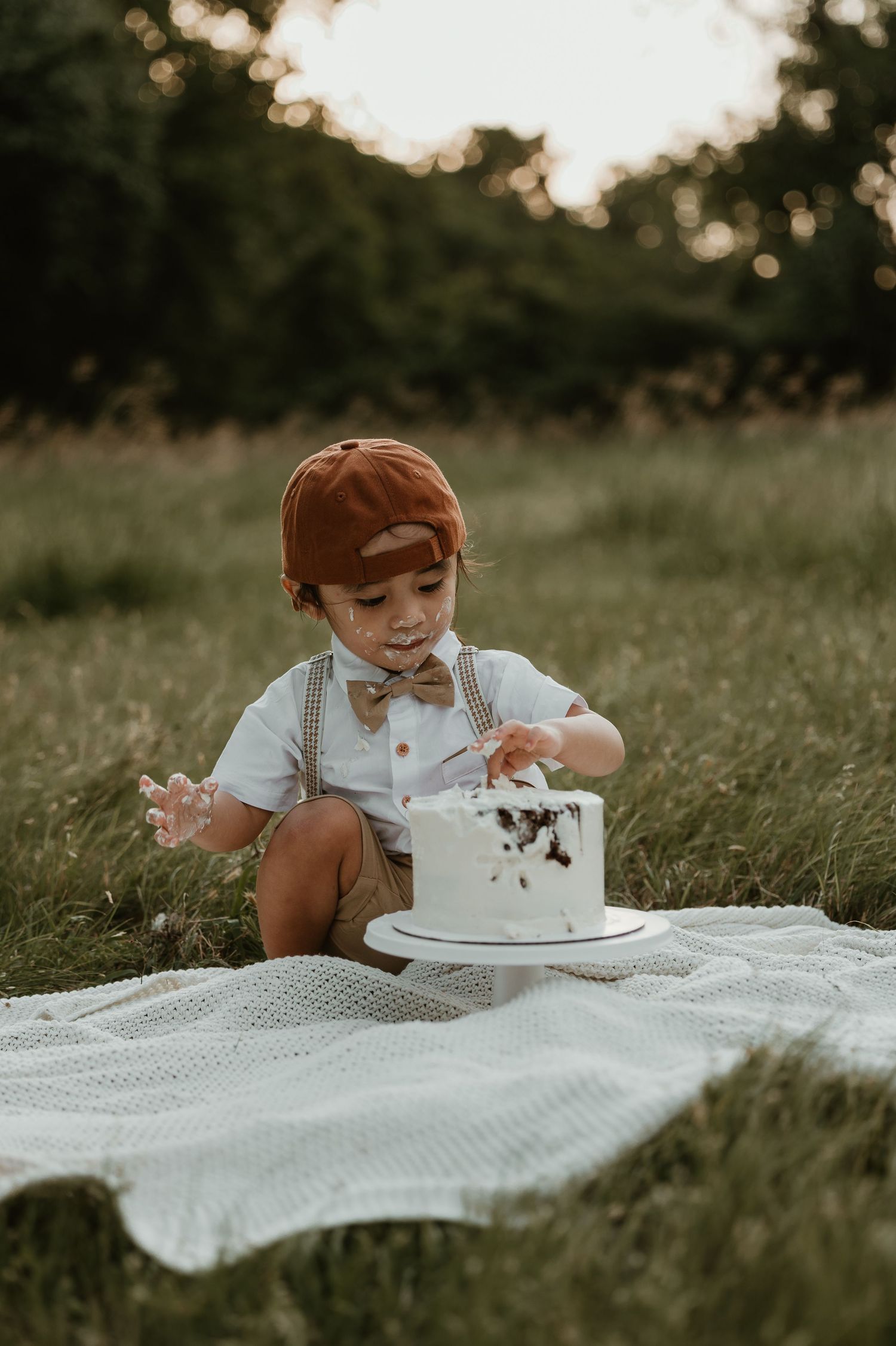 Small child in bow tie and suspenders playing with white cake on picnic blanket.