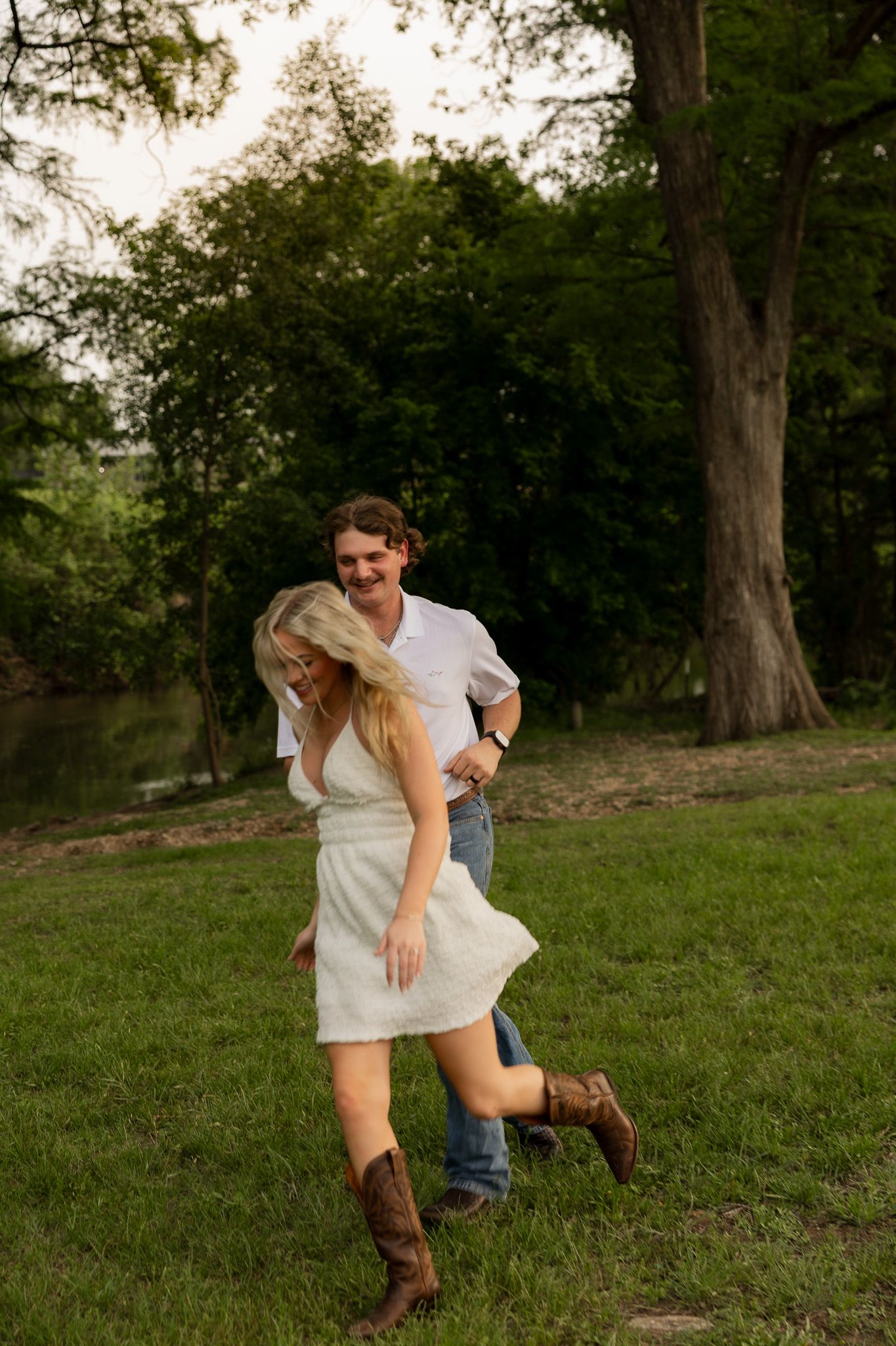 A joyful couple plays in a lush green park, woman in white dress and cowboy boots, man in white shirt and jeans.