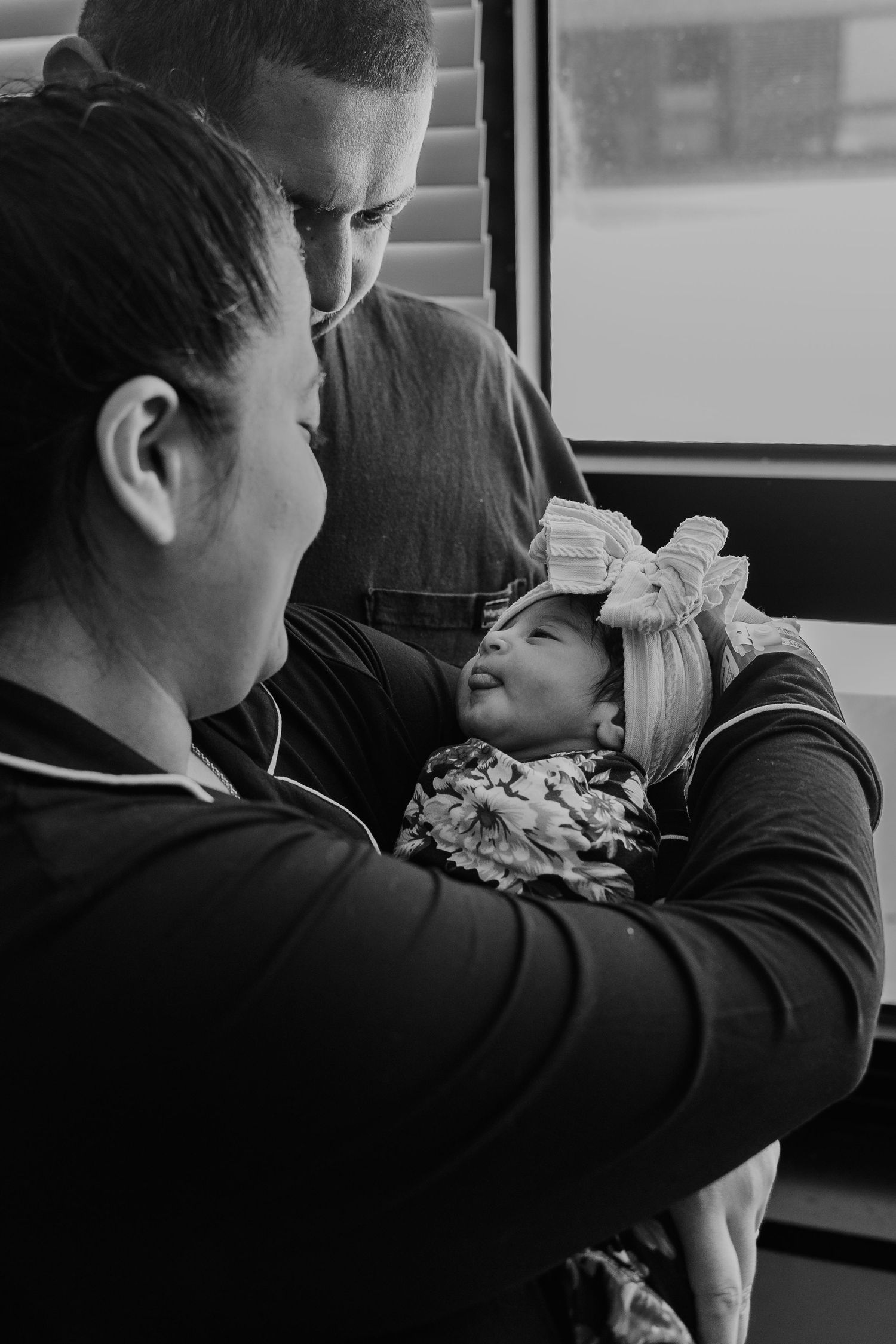 Black and white photo of parents lovingly gazing at their smiling newborn baby wearing a bow headband.