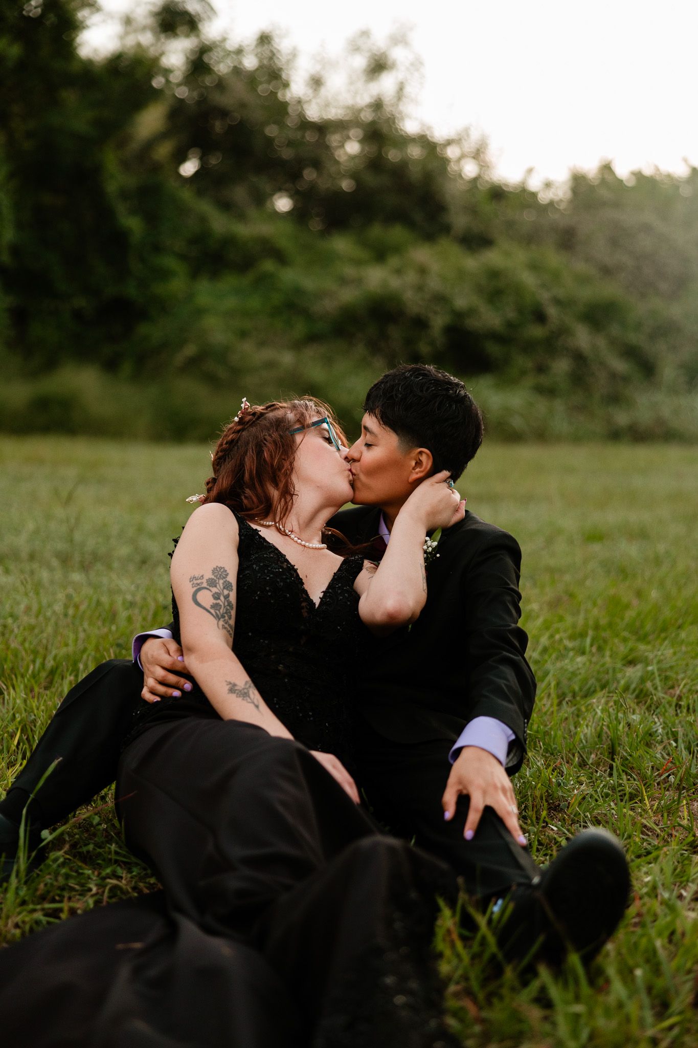 Couple in black formal wear sharing a romantic kiss while sitting in a lush green field at dusk.