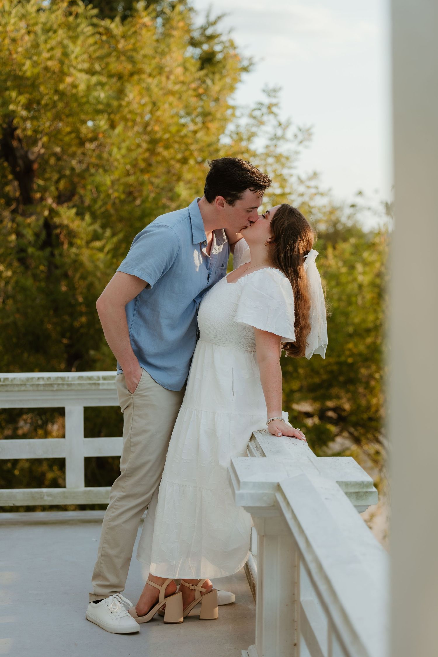 A couple shares a romantic moment on a white porch with autumn trees in background.