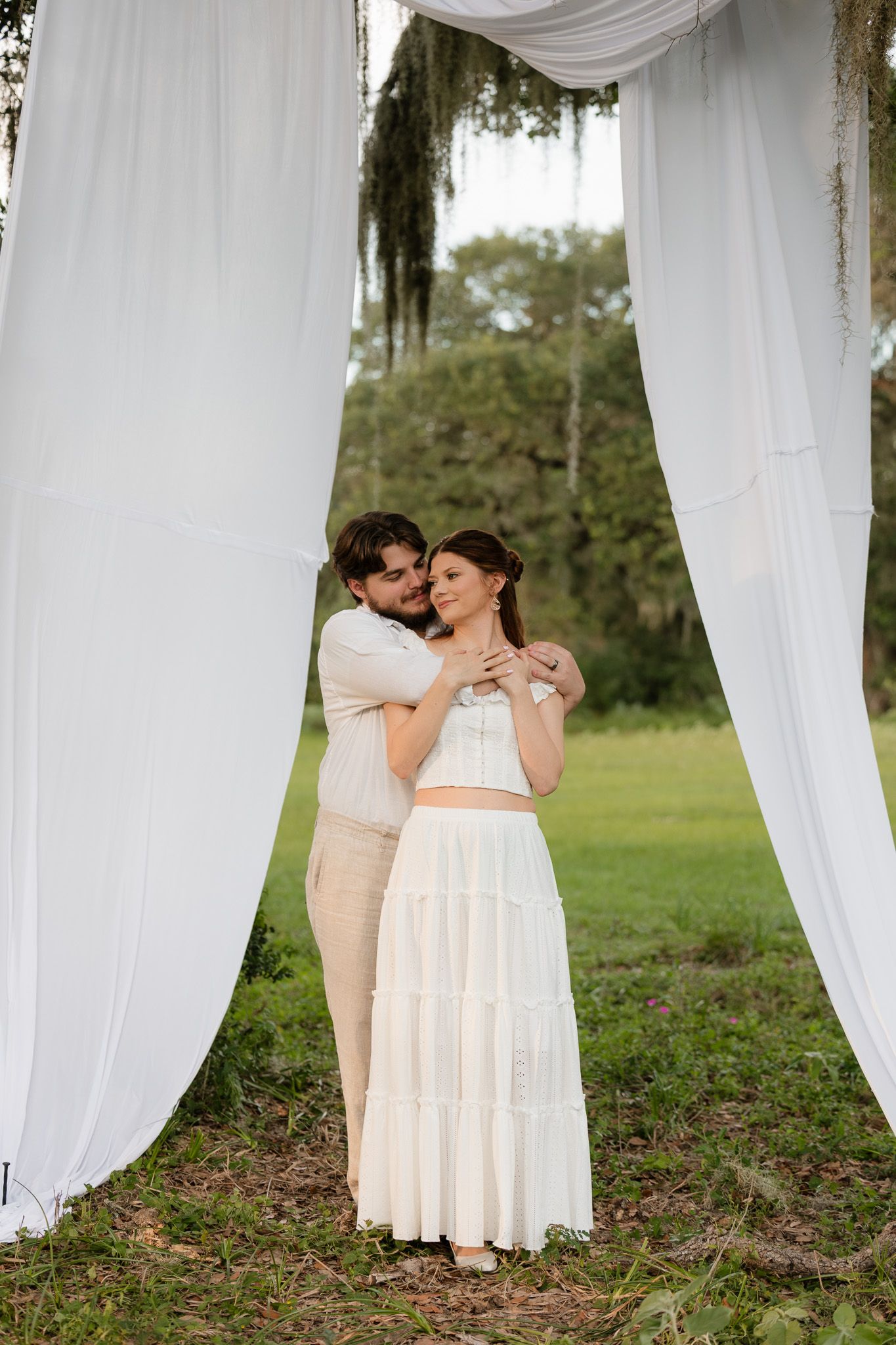 Couple embracing under white draping outdoors, woman in white tiered skirt and crop top, man in beige outfit, green field backdrop.
