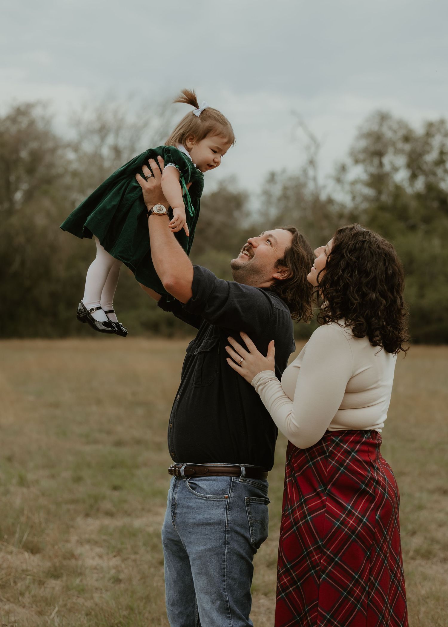 A joyful family outdoors, father lifting toddler girl in green dress while mother watches and smiles in a grassy field.