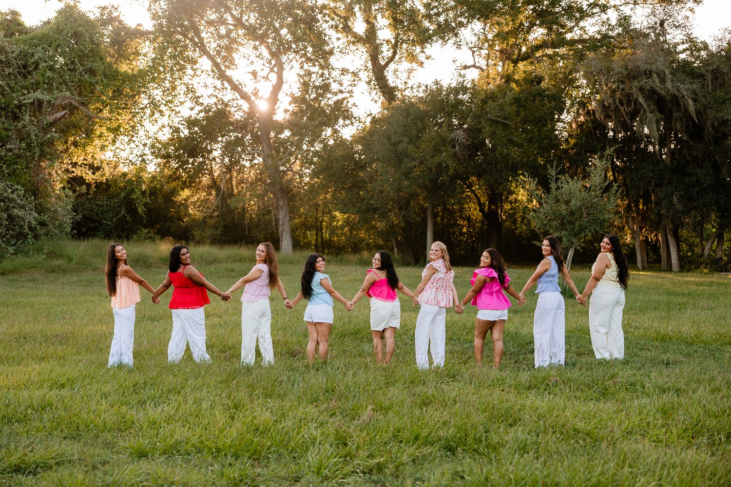 Eight women in colorful tops and white pants hold hands in a line on a sunlit green field surrounded by trees.