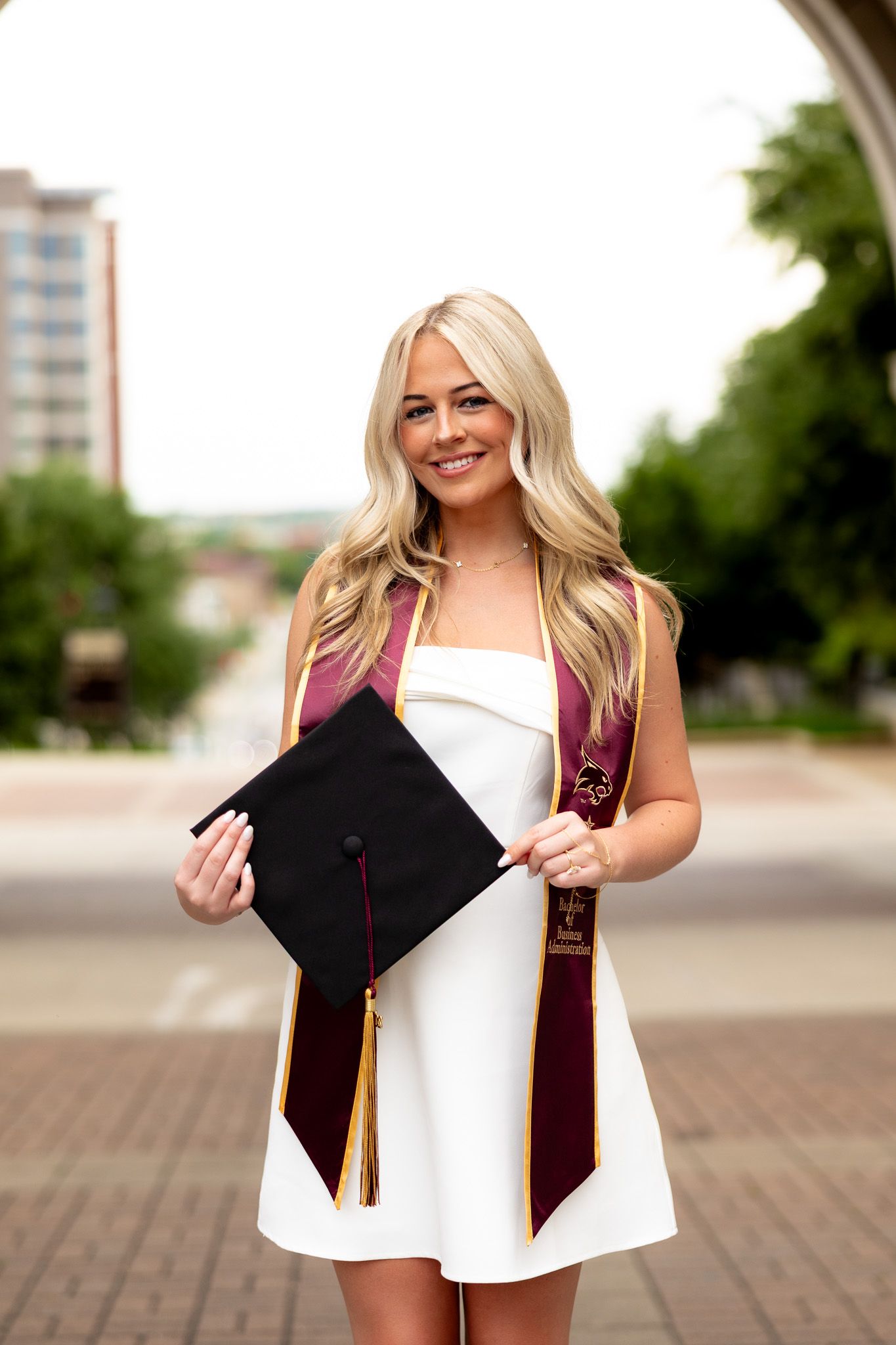 Smiling blonde woman in white dress holding graduation cap with maroon and gold stole outdoors.