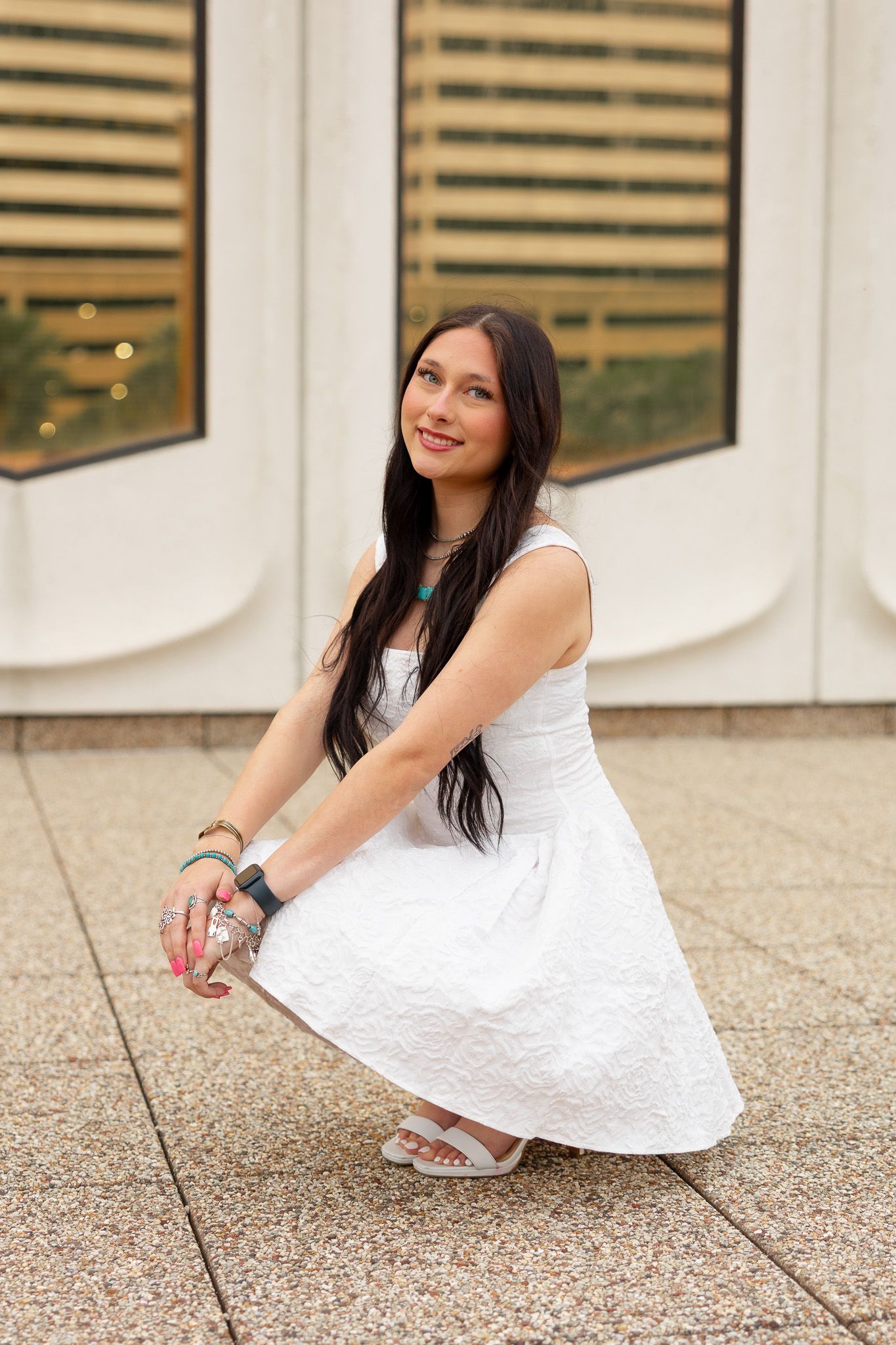 Young woman in white dress crouching outdoors, smiling, with dark hair and accessories, against a modern building.