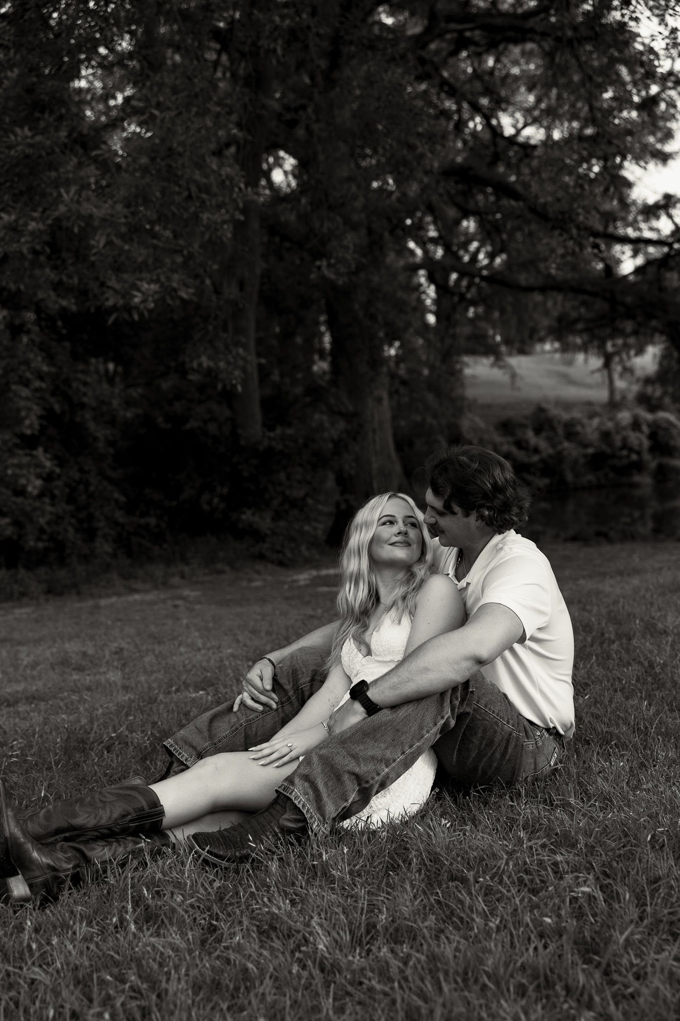 Black and white photo of a romantic couple sitting together on grass beneath large trees at dusk.