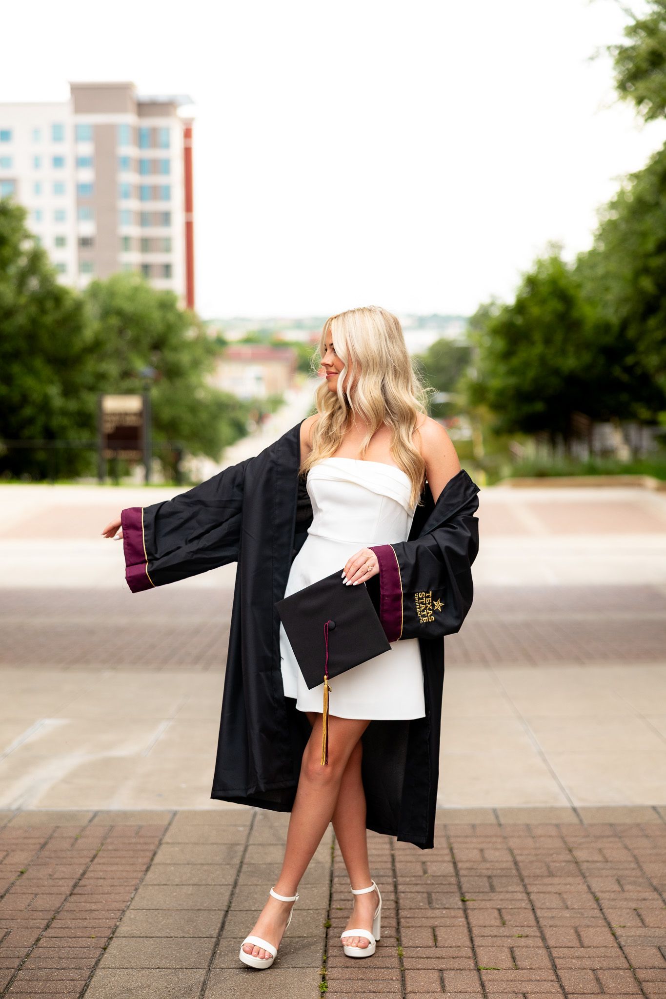 Blonde woman in white dress and black graduation gown holds cap outdoors on brick plaza with trees and buildings behind.