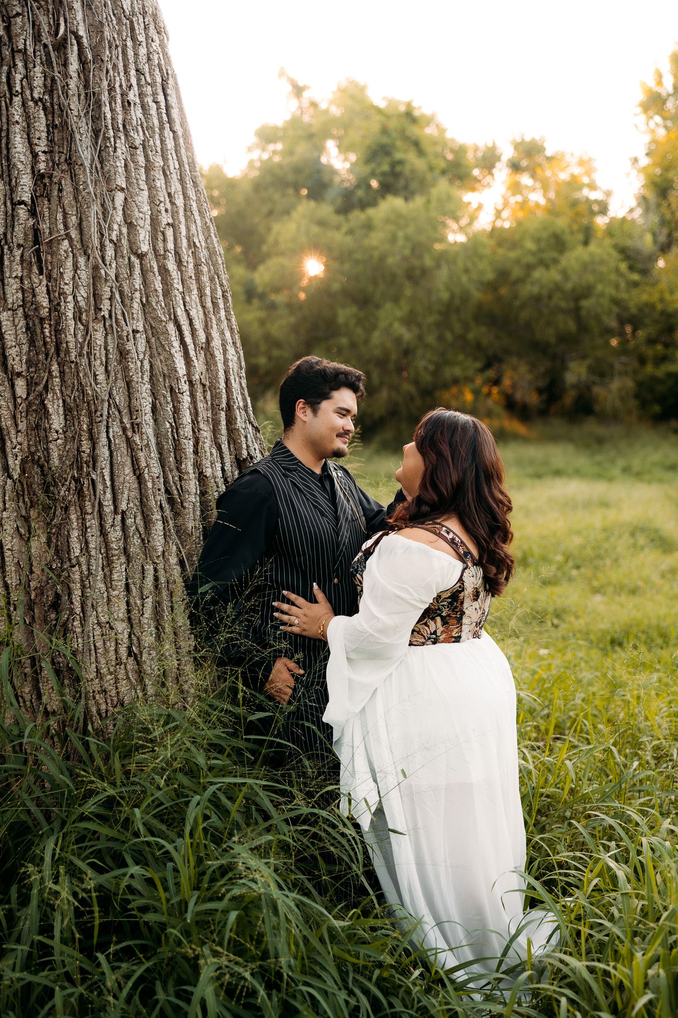 Couple sharing a romantic moment leaning against a large tree in a lush green field at golden hour.
