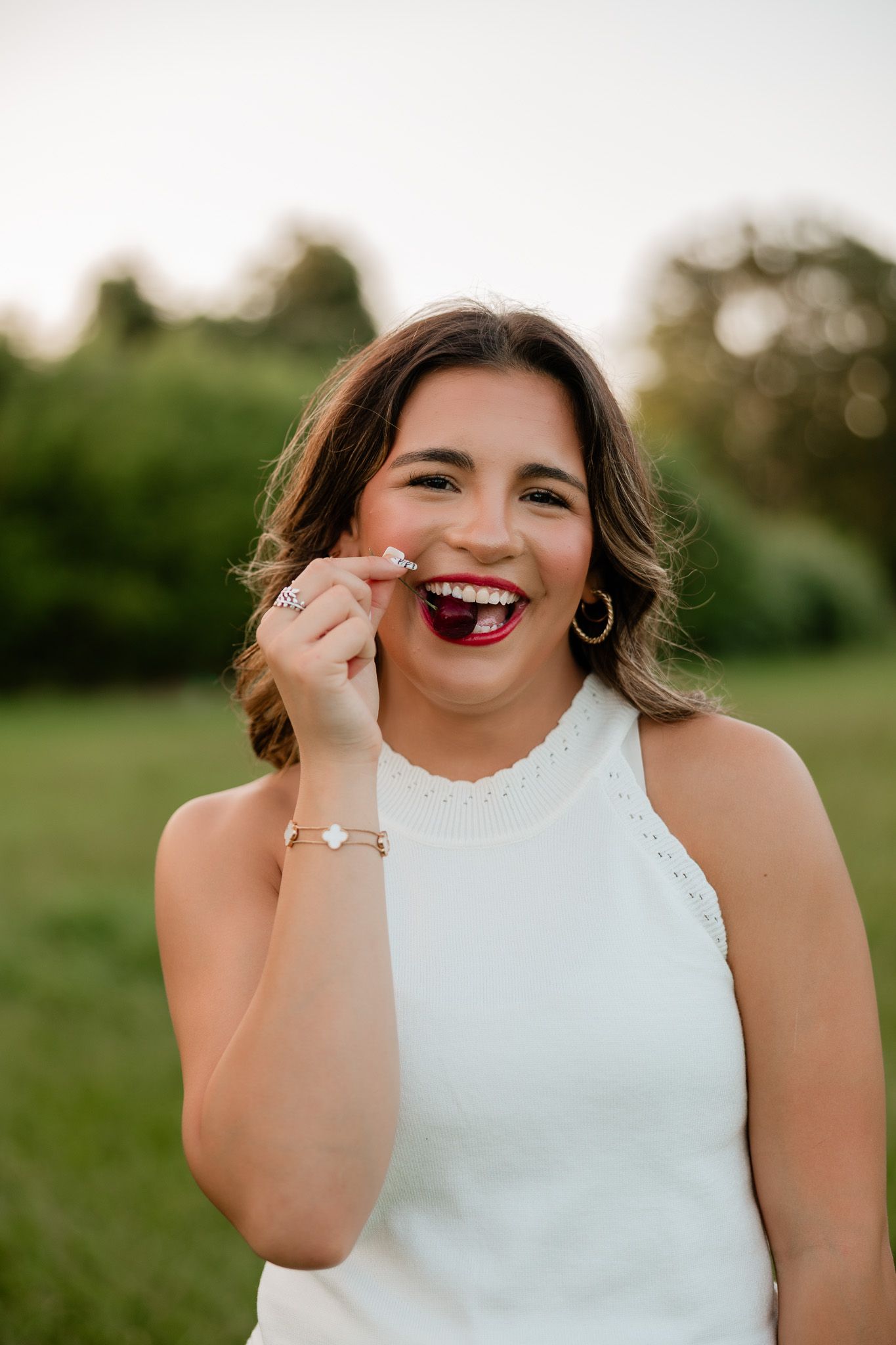 Smiling bride-to-be in white dress showing off engagement ring outdoors with green trees in background.