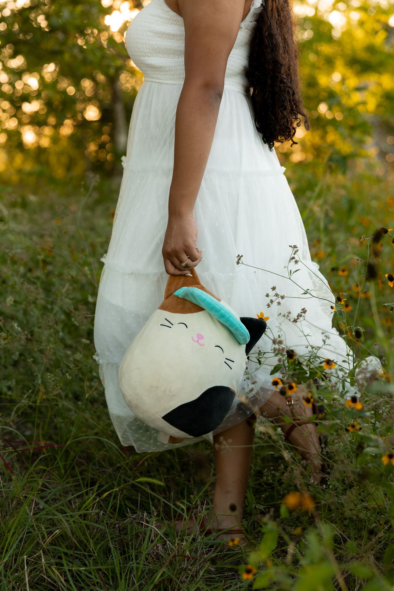 Girl in white dress holding a Squishmallow cat plush toy while walking through a wildflower meadow at golden hour.