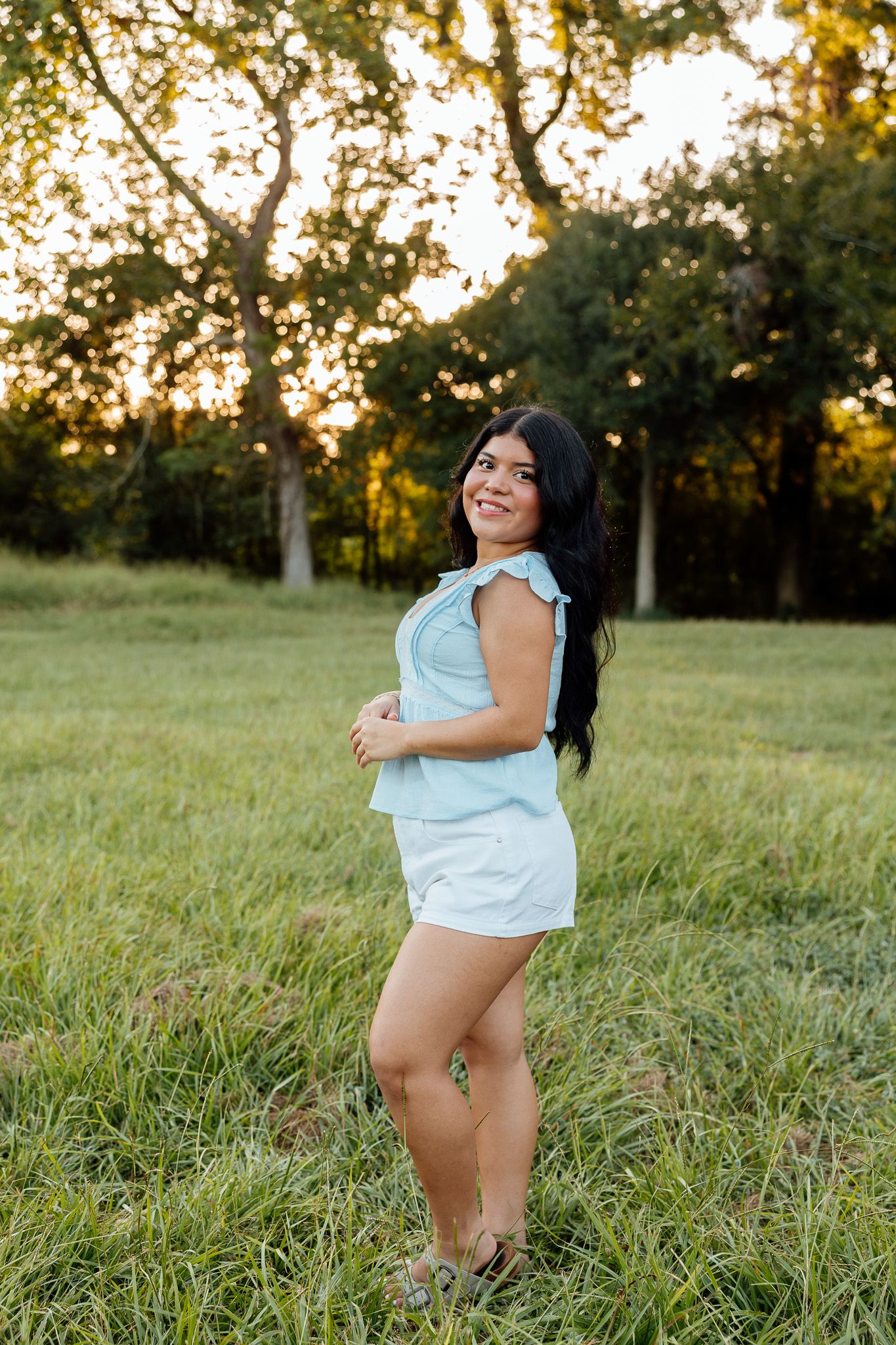 Young woman in light blue romper smiling in a grassy field at golden hour with trees in background.