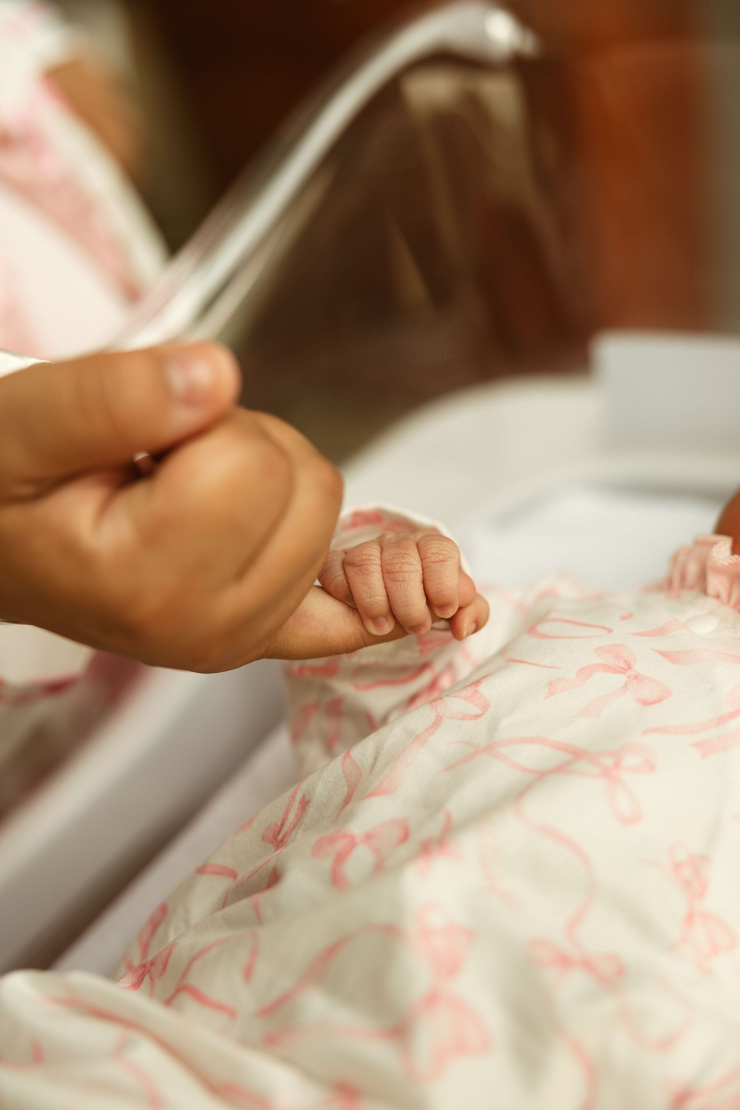 A parent gently holds a newborn baby's tiny hand in a hospital bassinet with white and pink patterned bedding.