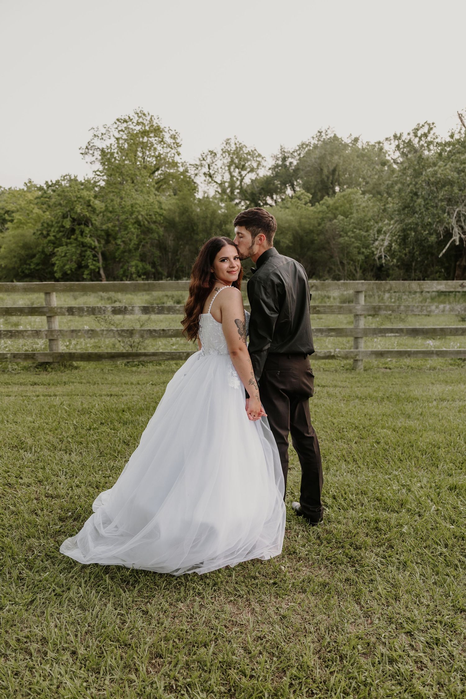 Bride in flowing white gown dances with groom in black suit on grass field.