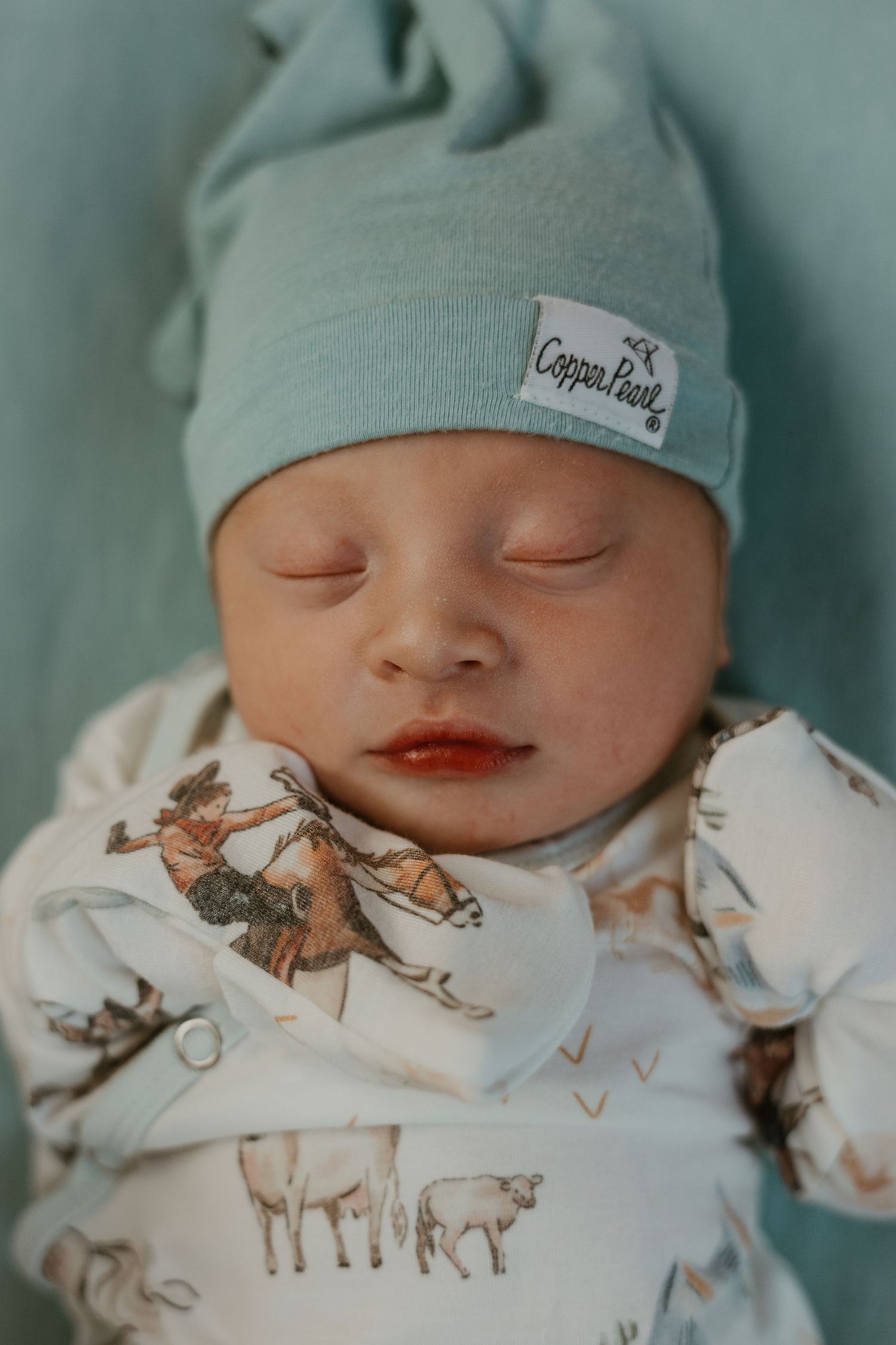 Sleeping newborn in mint green hat and animal print onesie.