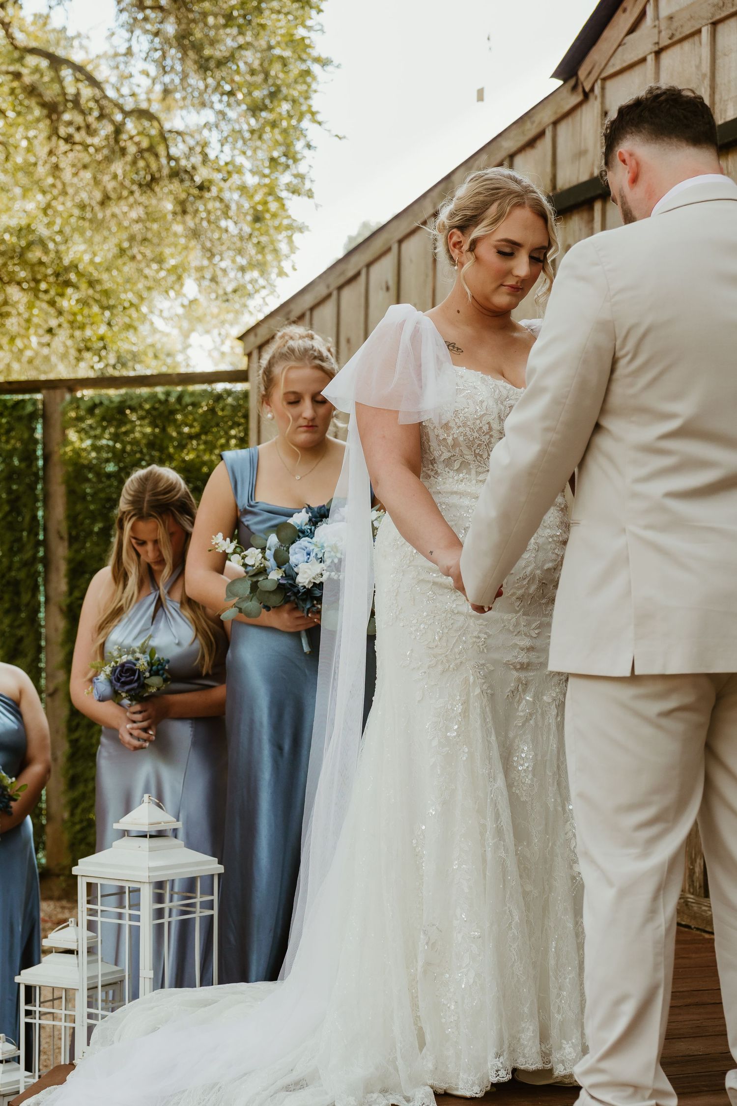 Outdoor wedding ceremony with bridesmaids in dusty blue dresses and white lantern decorations.