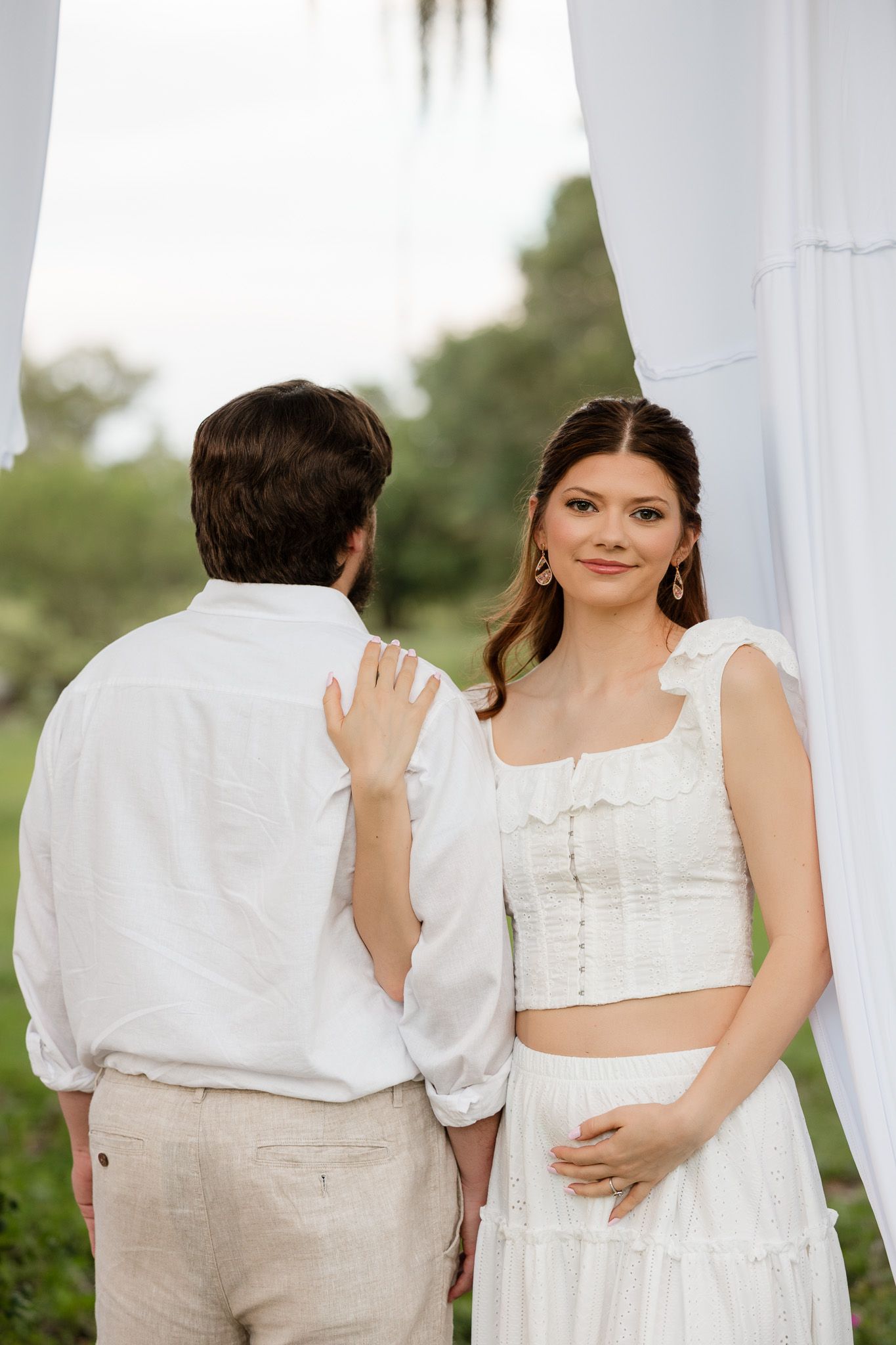 Couple in white outfits standing outdoors near white draping, woman smiling at camera during romantic outdoor event.