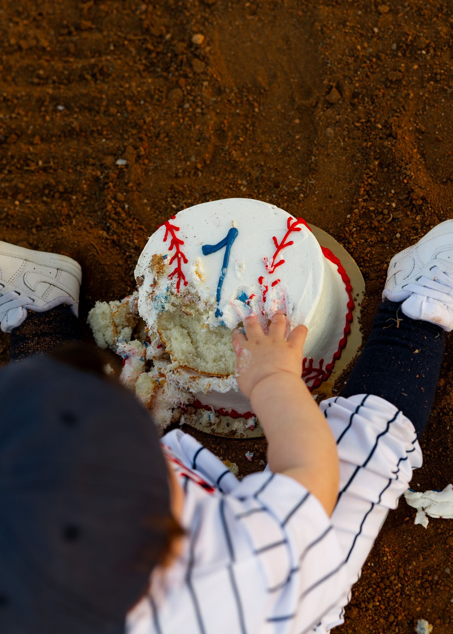Baby in baseball uniform smashing their first birthday baseball cake on a dirt field.