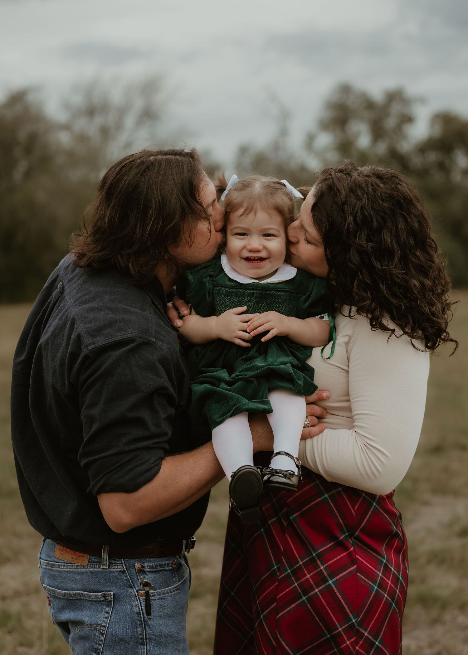 Parents kissing cheeks of smiling toddler girl in green dress during outdoor family portrait session.
