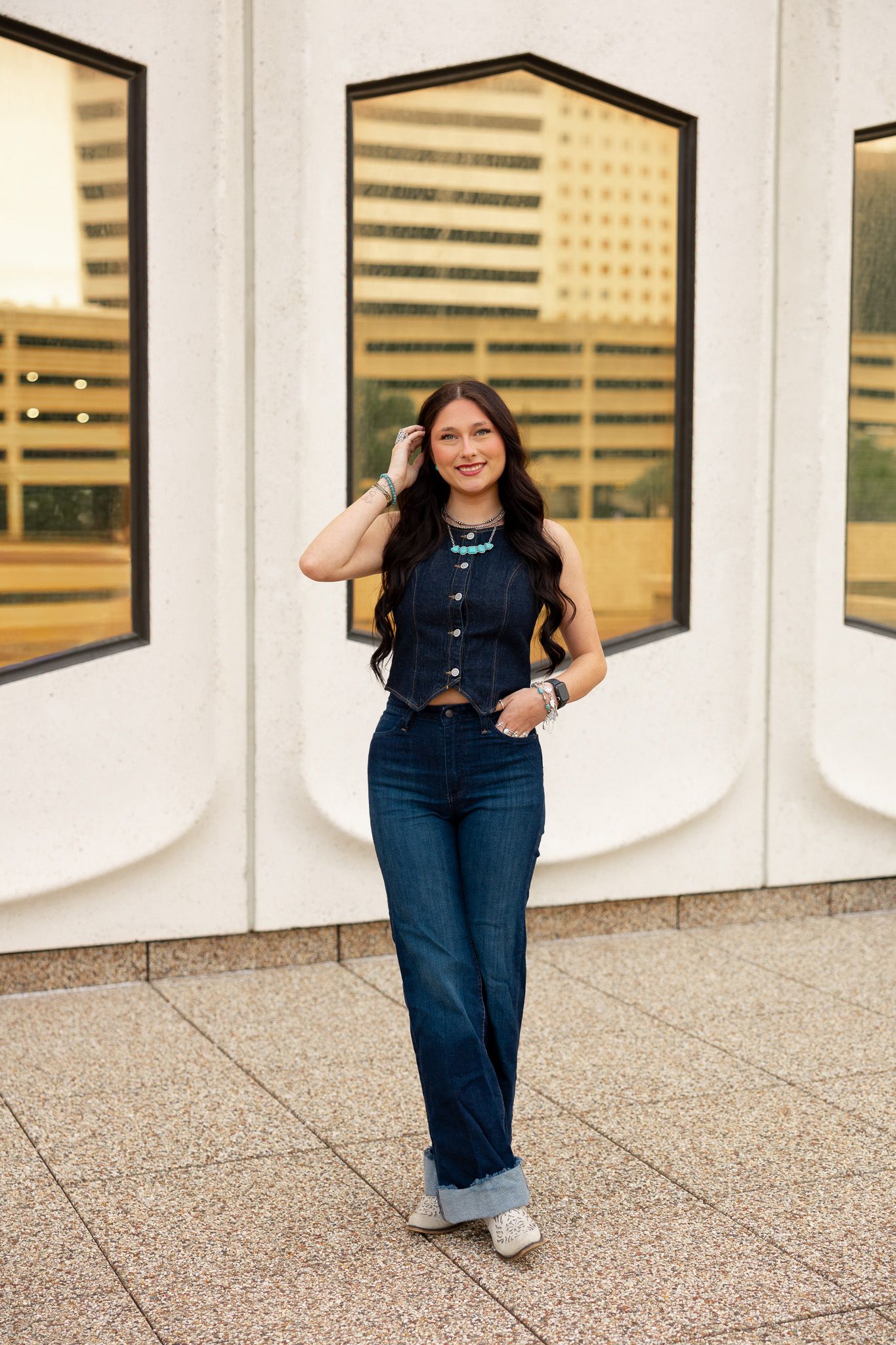 Young woman with long dark hair in dark denim outfit wearing turquoise necklace poses outdoors.