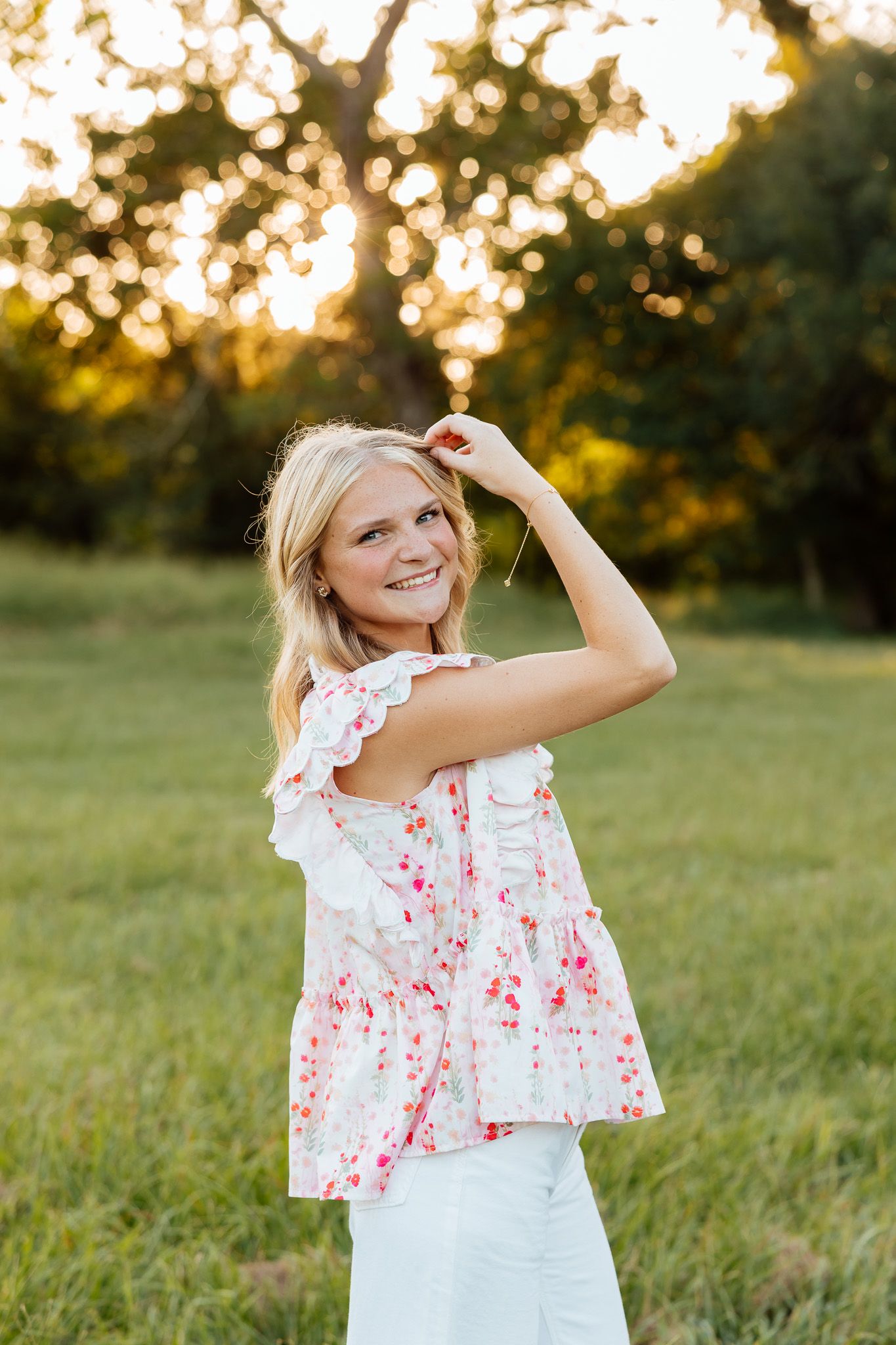 Young blonde woman in floral top smiling outdoors at golden hour in a green field.