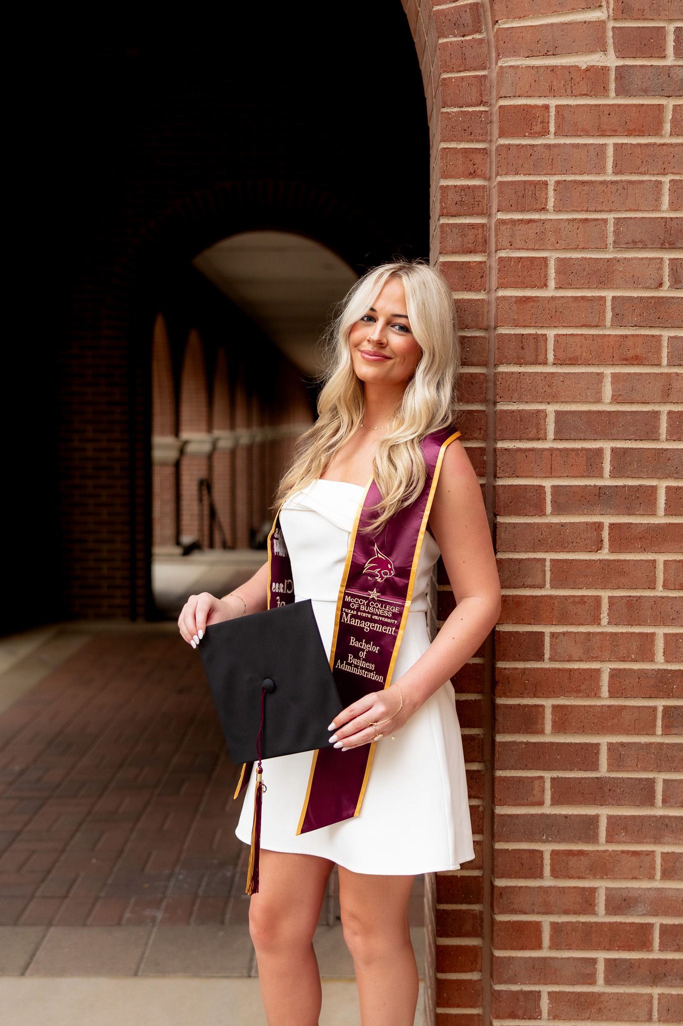Blonde woman in white dress holding graduation cap wearing maroon sash poses by brick wall at university.