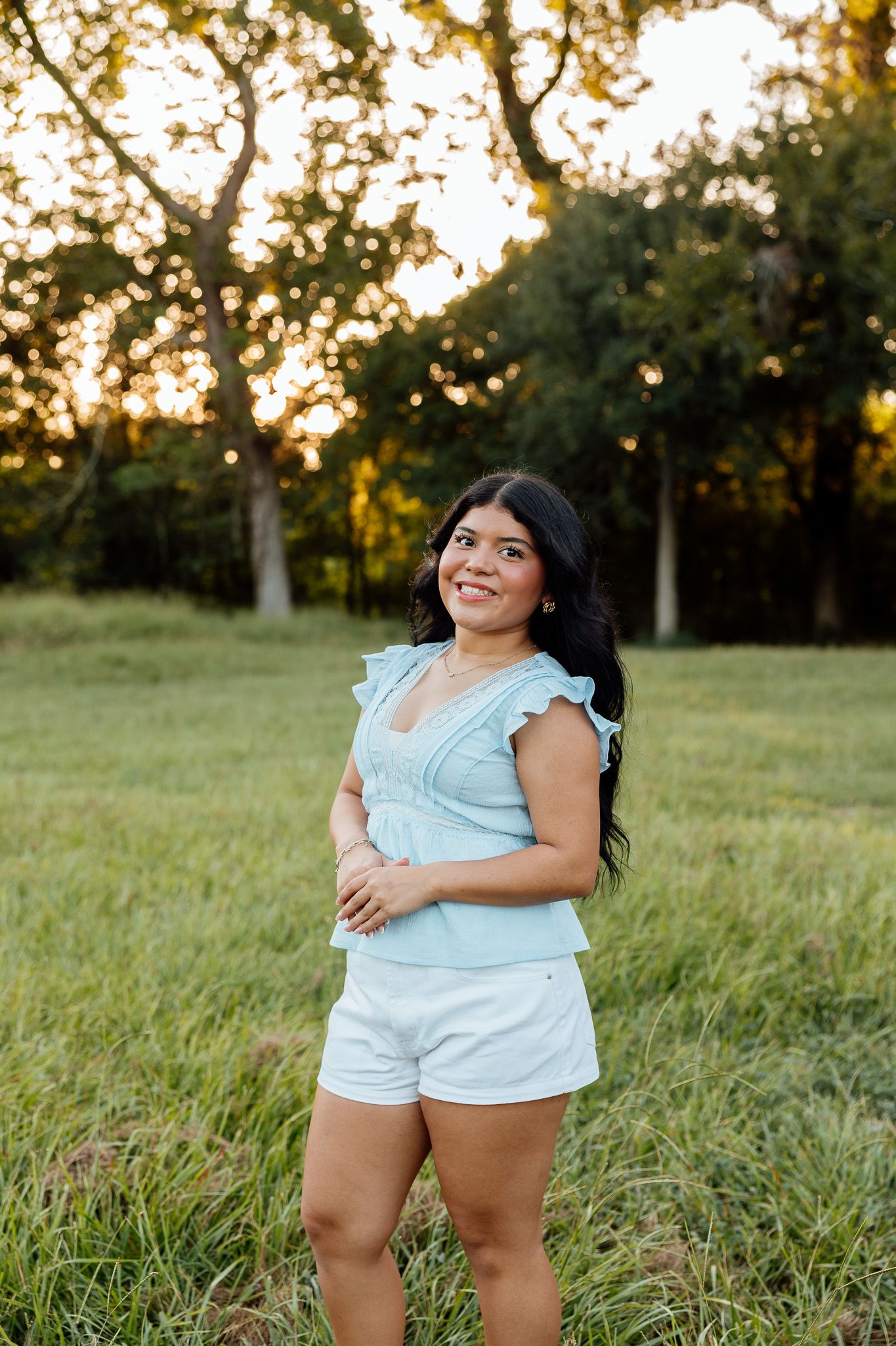 Young woman in light blue top and white shorts smiling outdoors in a grassy field at golden hour.