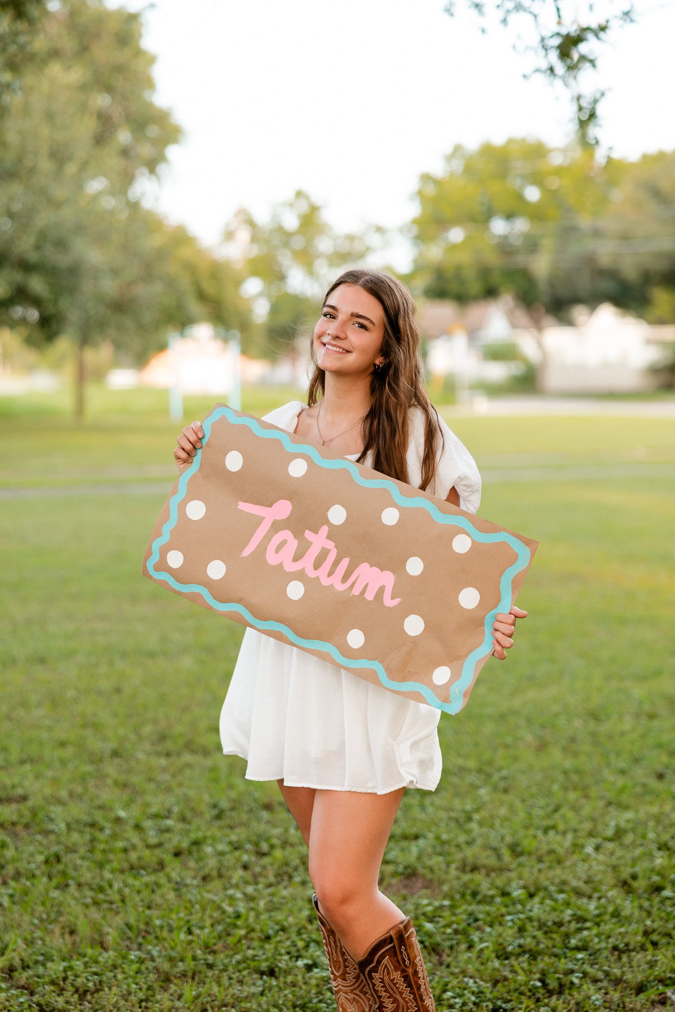 Young woman in white dress and cowboy boots holding a decorative burlap sign reading Tatum outdoors.