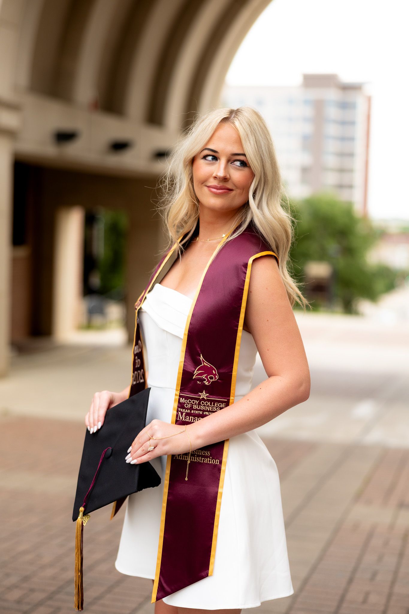 Blonde woman in white dress wearing maroon graduation stole holds cap outdoors at university campus.